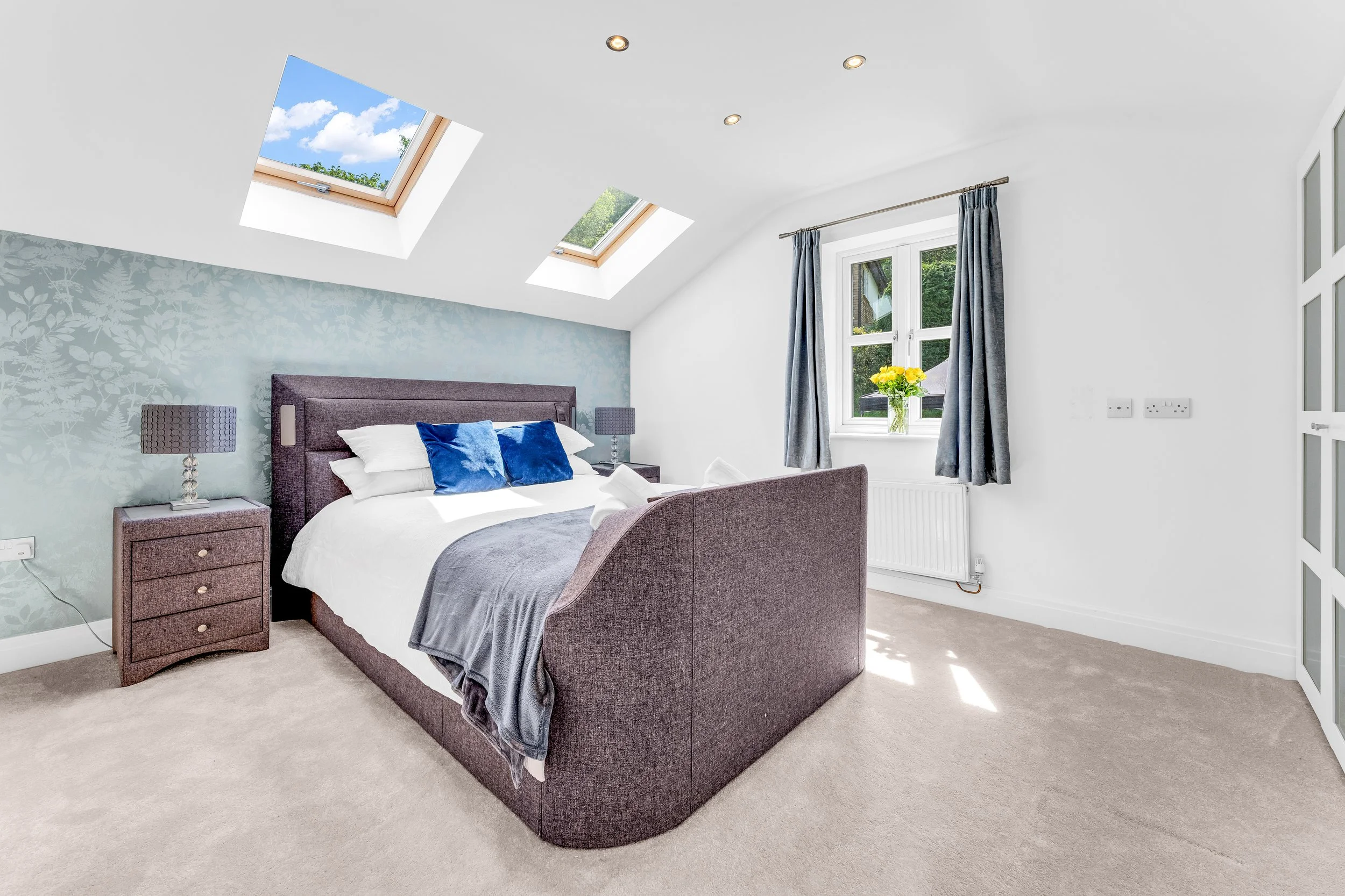 A bedroom with a bed featuring a dark gray upholstered headboard, white bedding, and blue pillows. Two matching gray bedside lamps sit on nightstands. The room has two skylights on the ceiling and a window with blue curtains, with a vase of yellow fl