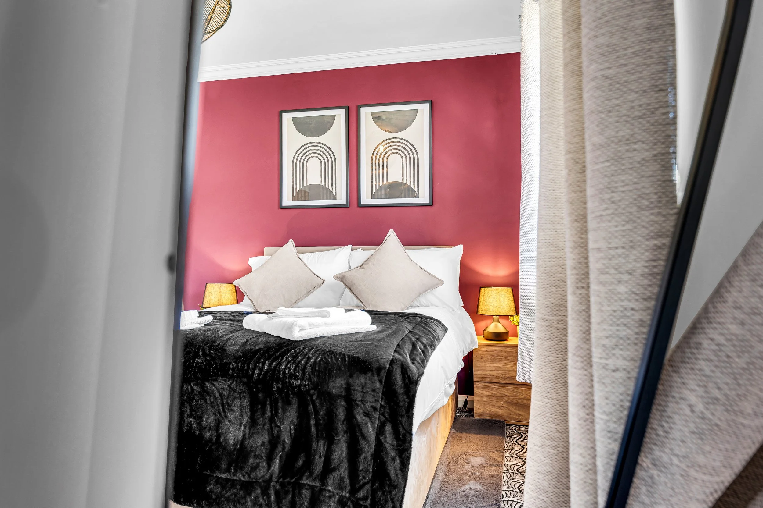 A neatly made bed with white pillows and beige cushions in a bedroom with red accent wall, framed abstract artwork, two bedside lamps, beige curtains, and a mirror reflecting the room.