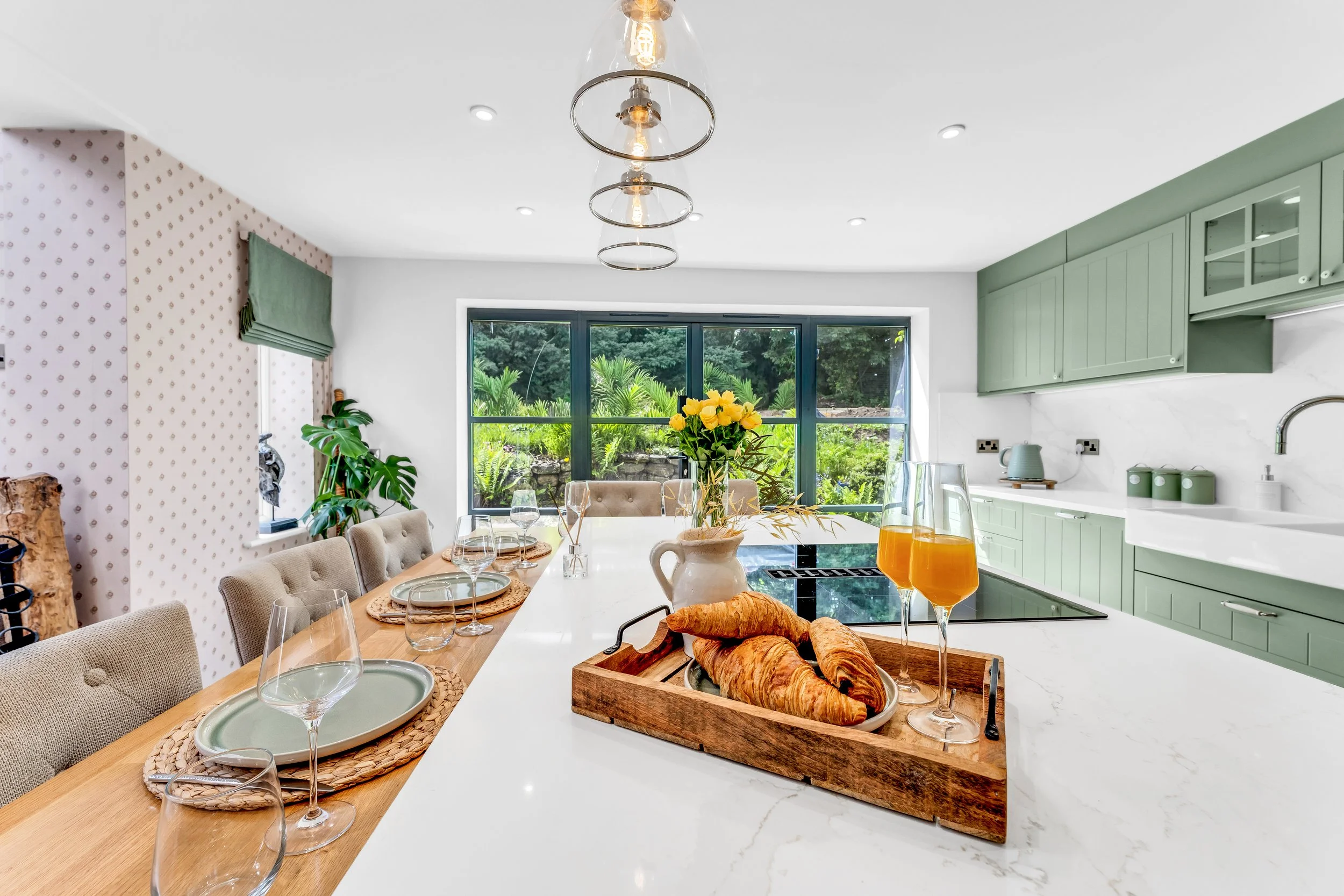 A bright dining area with a large window showing a garden outside. The table is set with plates, glasses, and a centerpiece of yellow flowers. Croissants and orange juice are on the table. The kitchen has green cabinets and a white marble countertop.