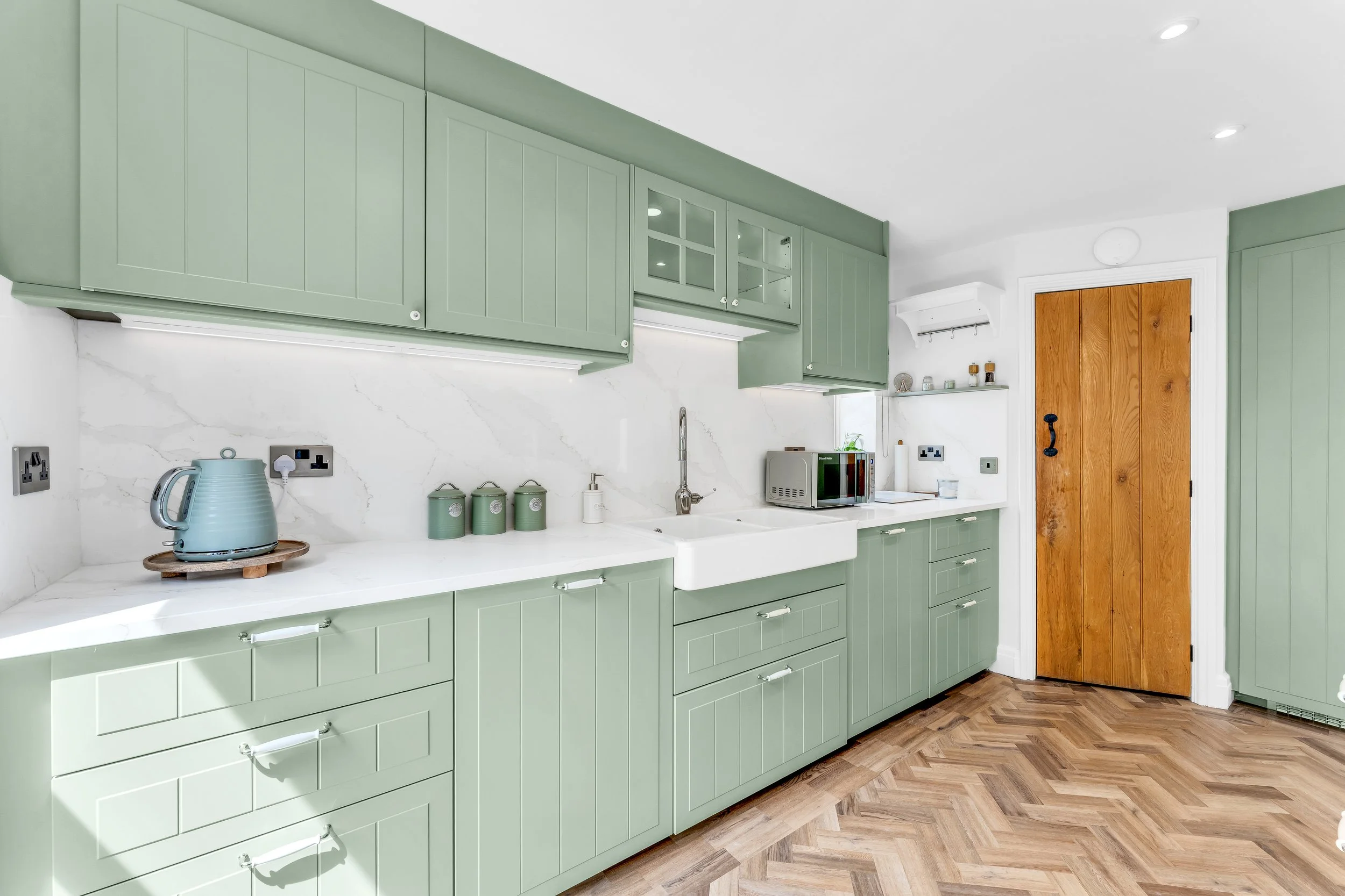 A modern kitchen with green cabinets, a white marble backsplash, and wooden flooring. There are kitchen appliances and decorative items on the countertops, including a kettle, jars, and a microwave. A wooden door is visible on the right.