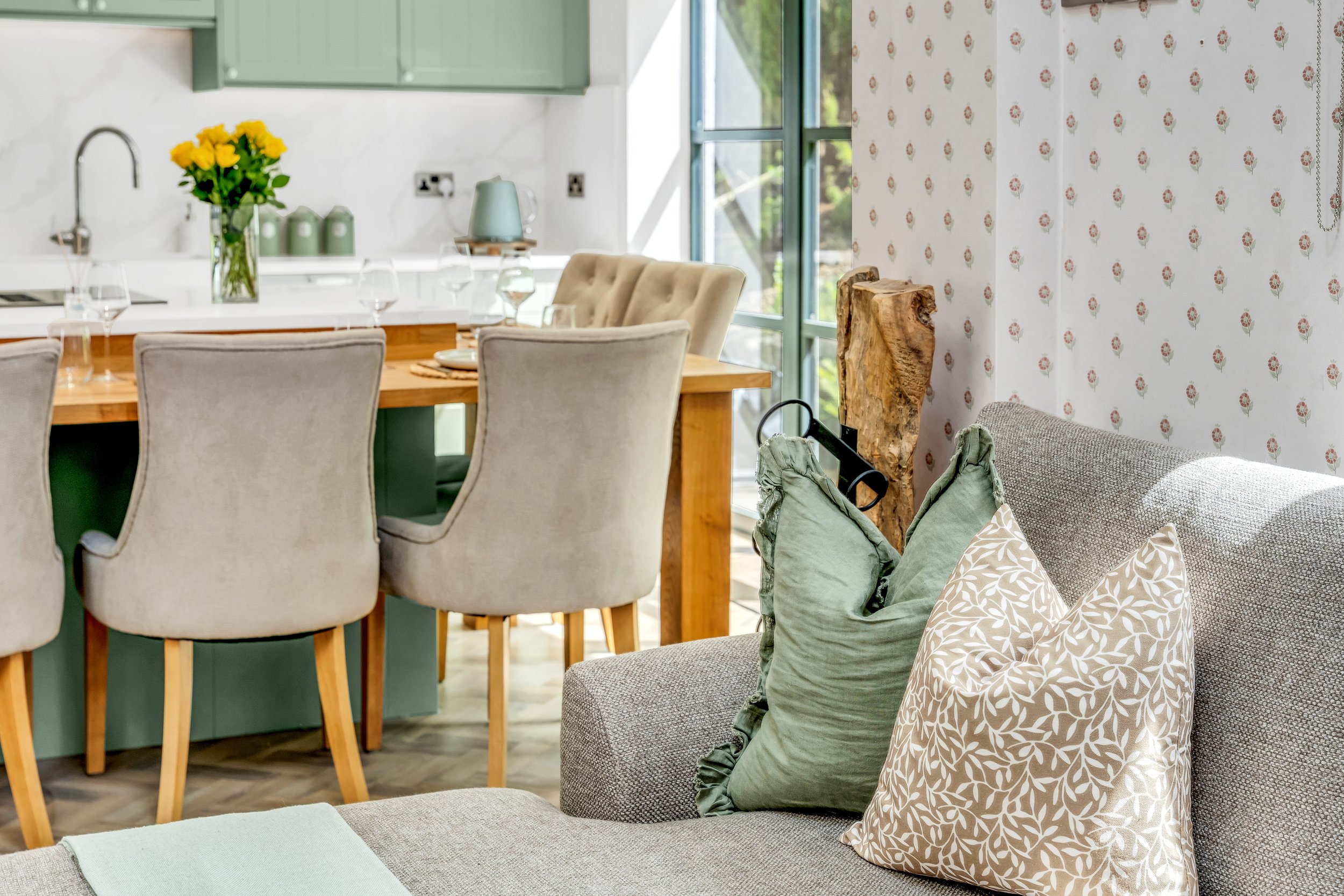 Living room with beige sofa and pillows, wooden table with chairs, large window with greenery outside, patterned wallpaper, and kitchen area with a vase of yellow flowers.
