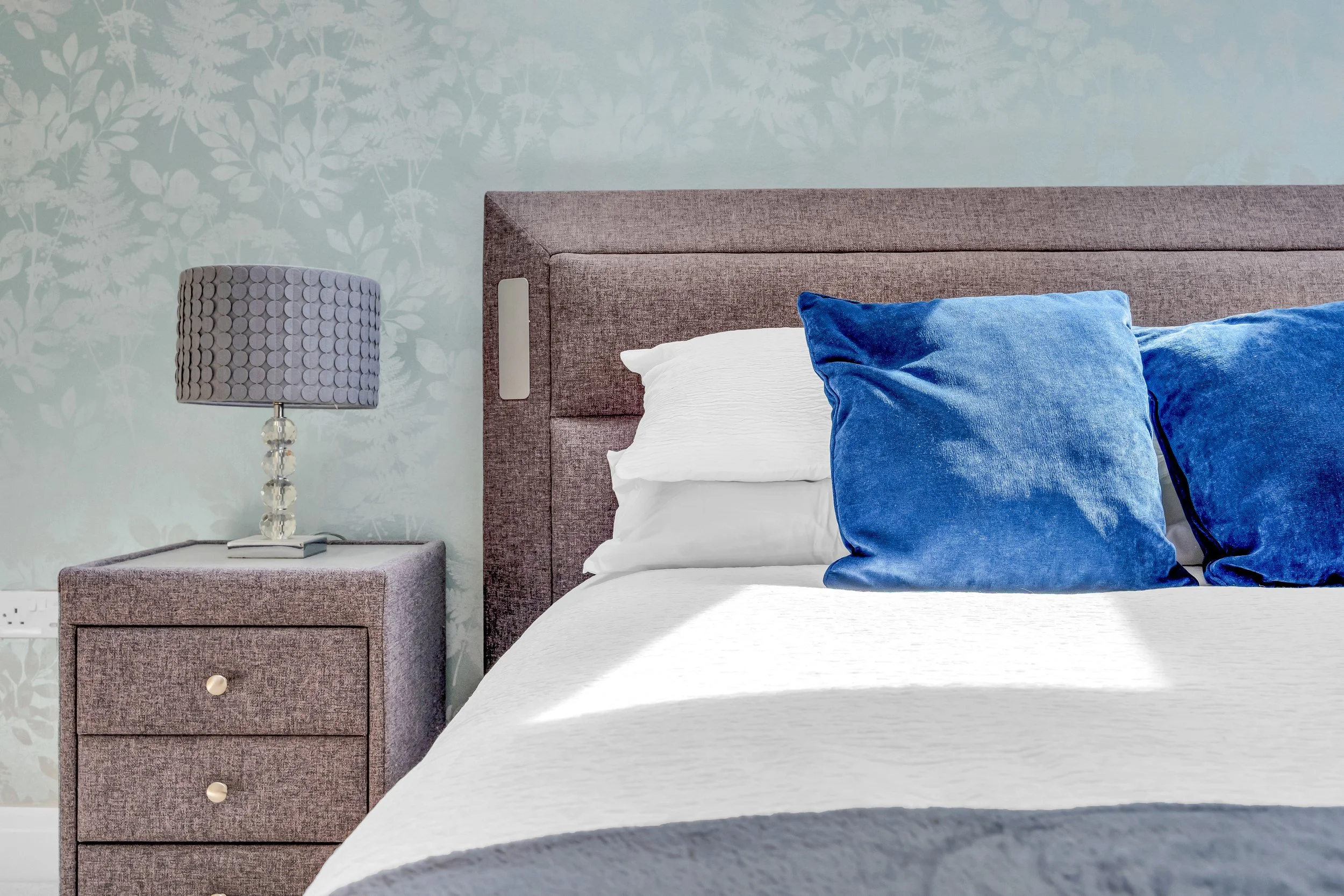 A bedroom with a gray upholstered bed head, white bedding, and two blue velvet pillows. There is a matching gray bedside table with a decorative lamp and a book on top. The wall behind has a light teal wallpaper with a leafy pattern.