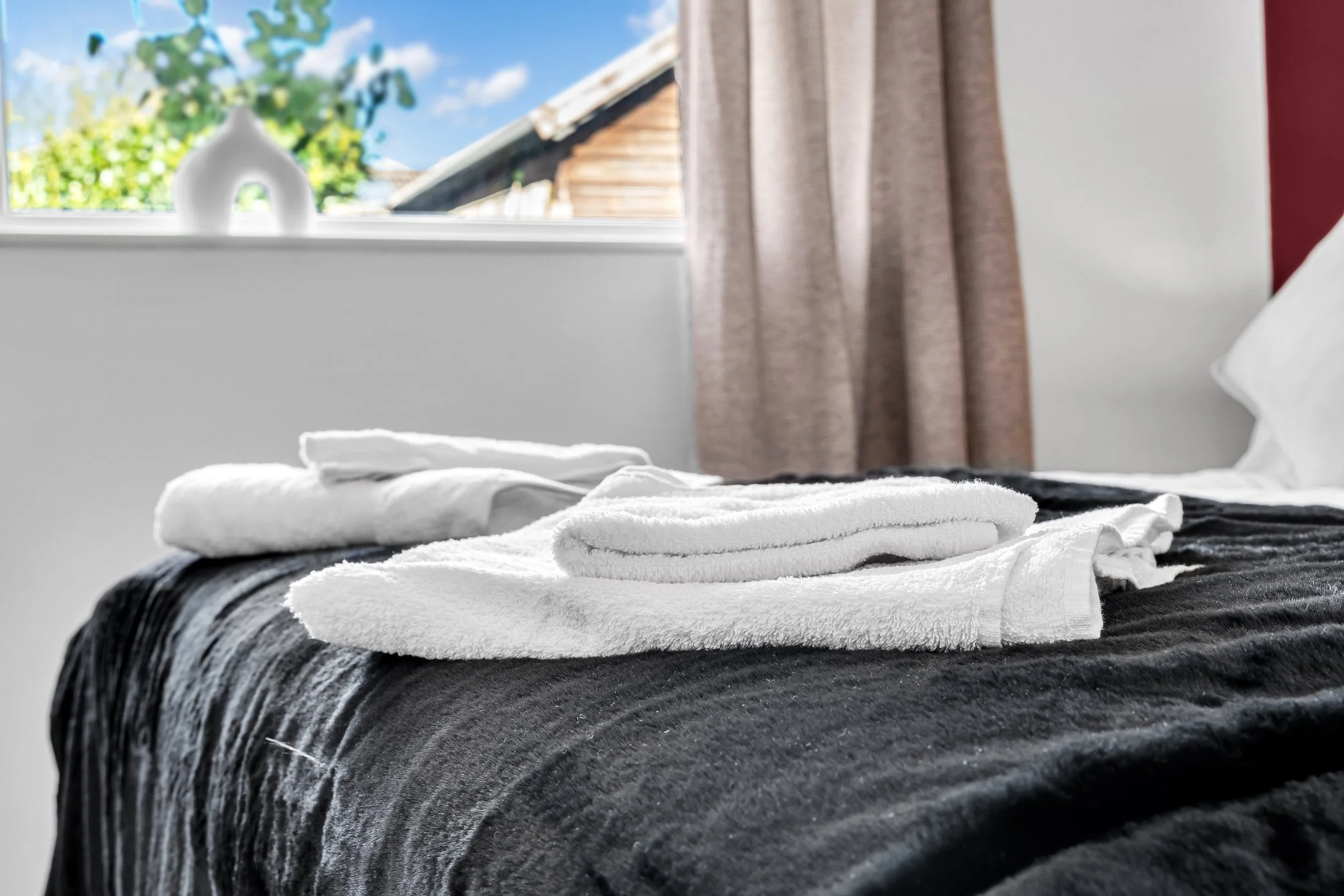 Towels folded on a bed with a black and gray striped blanket, near a window with beige curtains, in a bedroom with wood siding visible outside.