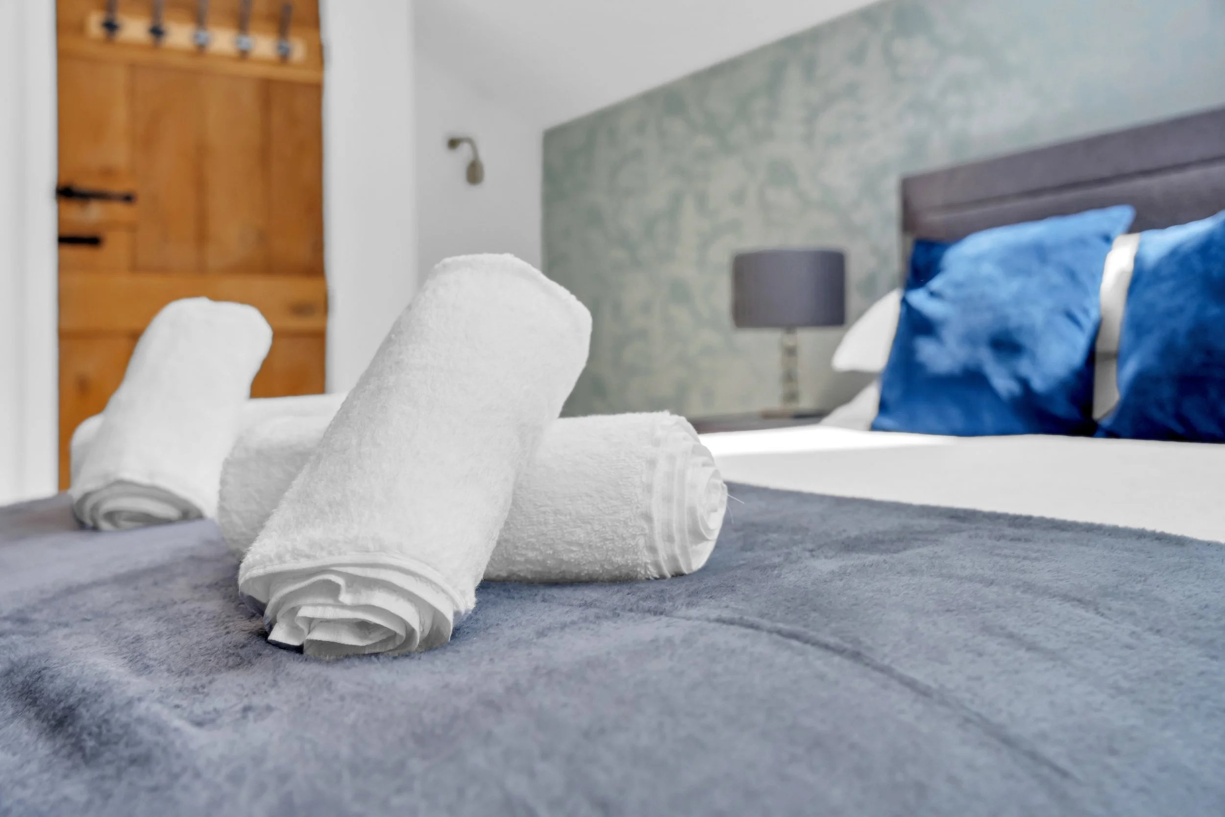 A hotel room with a bed that has blue pillows, a wooden headboard, and a blue and gray patterned wall. In the foreground, there are three neatly rolled white towels on a gray carpeted floor, with a small white towel underneath each rolled towel.