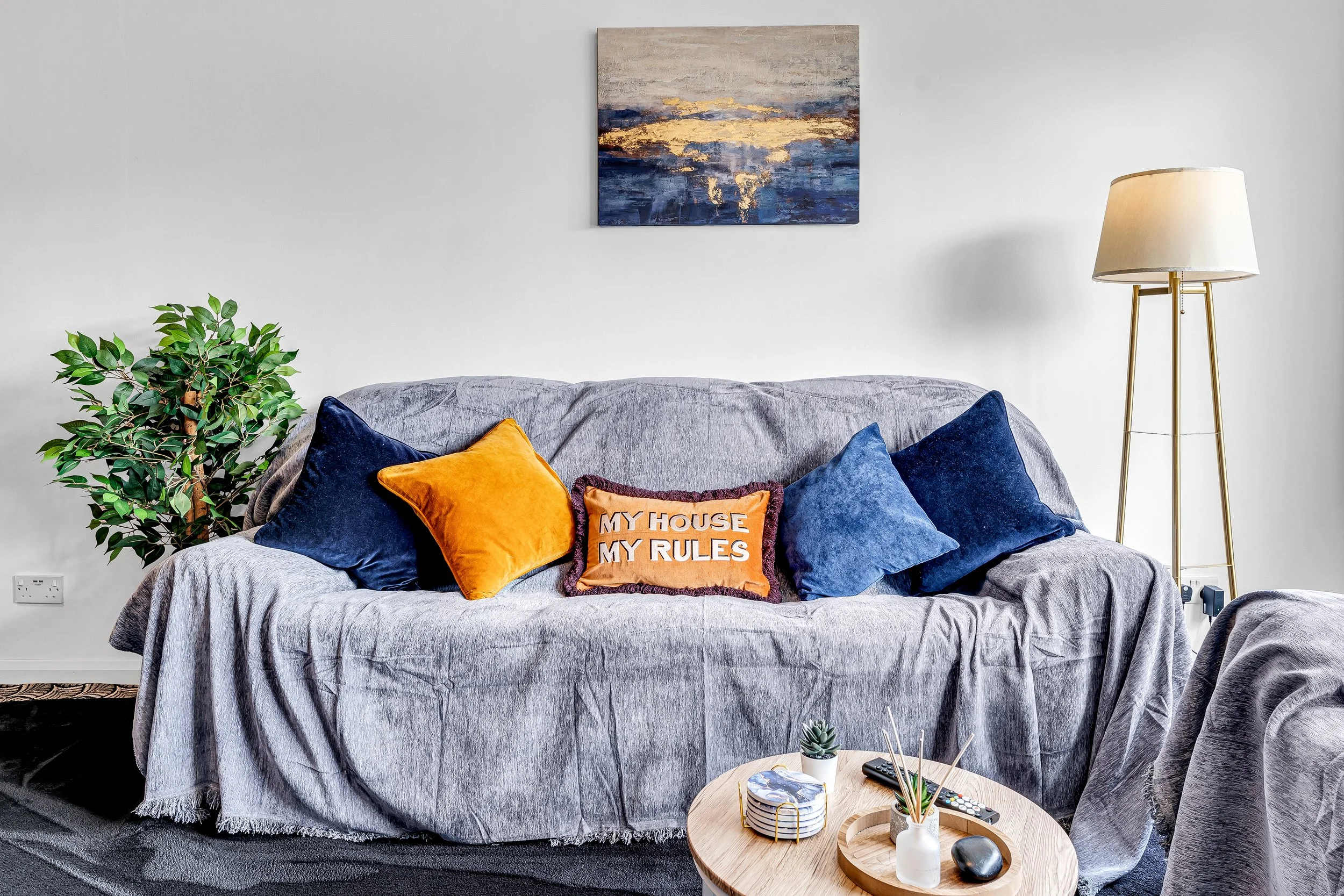 Living room with a gray sofa covered with a blanket, topped with five pillows including a decorative pillow that says 'My House My Rules,' a small circular wooden coffee table with a small plant, magazines, incense, and remote control, a potted green