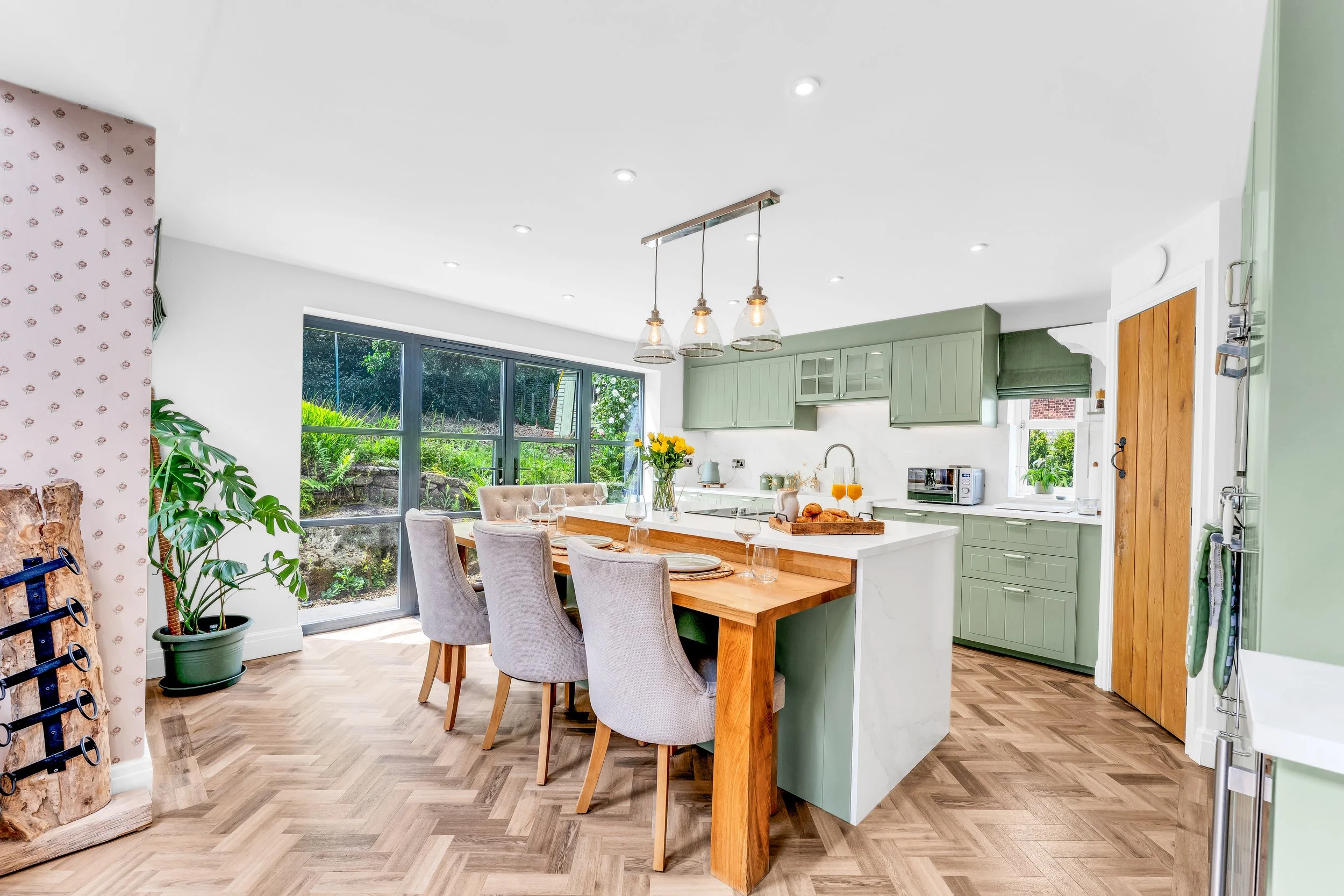 Bright kitchen and dining area with green cabinets, a large window, and a wooden dining table with beige chairs. Decorations include a potted plant, a bouquet of yellow flowers, and a tray of croissants on the table.