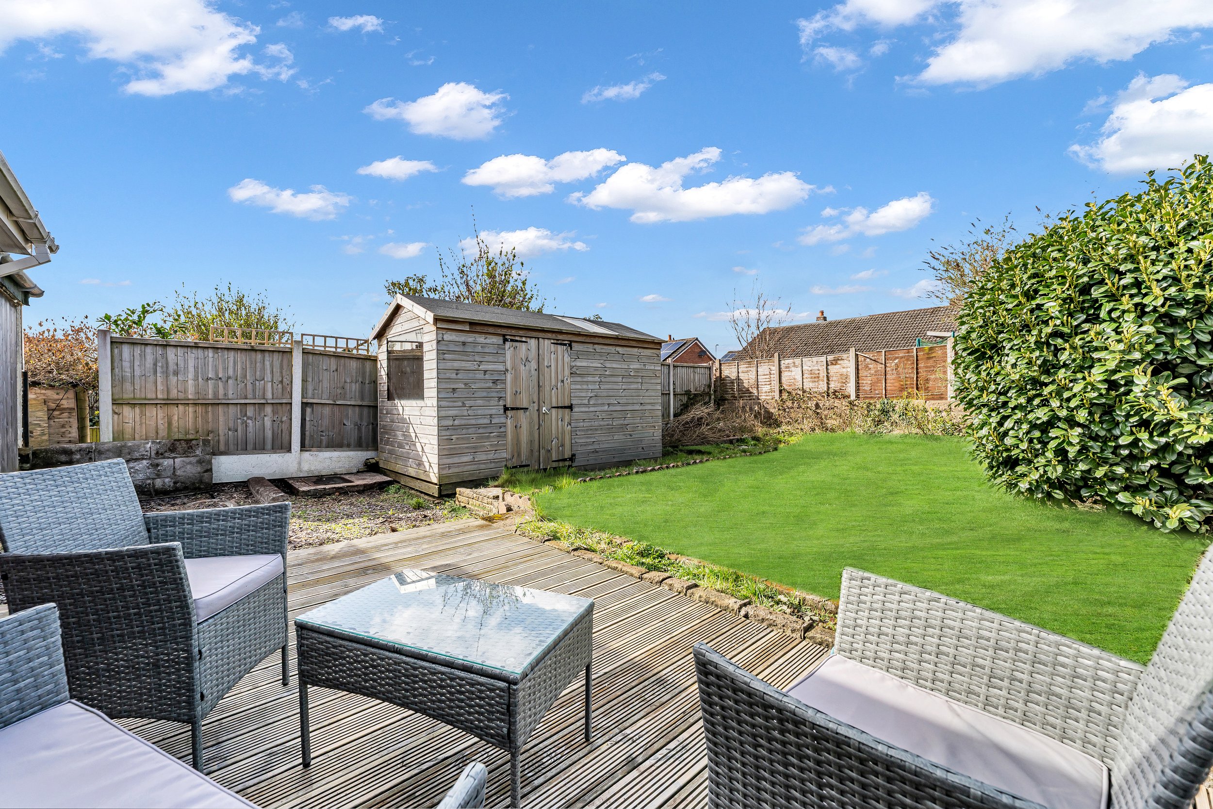 A backyard with a wooden deck, outdoor furniture including chairs and a glass-topped table, a well-maintained lawn, a wooden shed, and a fence under a bright blue sky with scattered clouds.