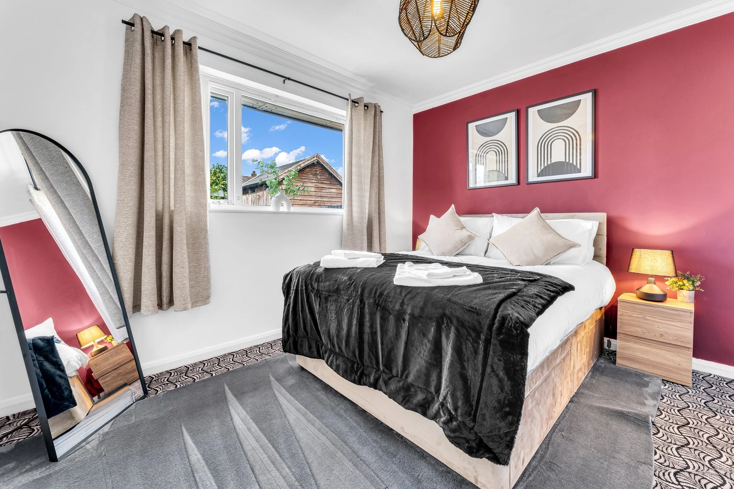 Bedroom with a double bed, pink accent wall, two framed abstract art pieces, beige curtains, window with a view of a wooden building and blue sky, full-length mirror, bedside lamp, small plant, black blanket on bed, neatly folded towels, patterned ca
