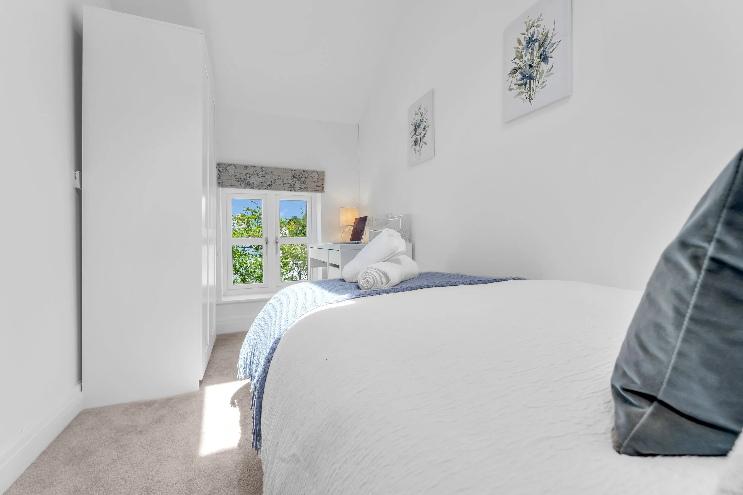 A white bedroom with a neatly made bed, decorative pillows, a rolled towel, and a blue and white striped blanket. There are two paintings of flowers on the wall and a window with a Roman shade displaying trees outside. A bedside table with a lamp and