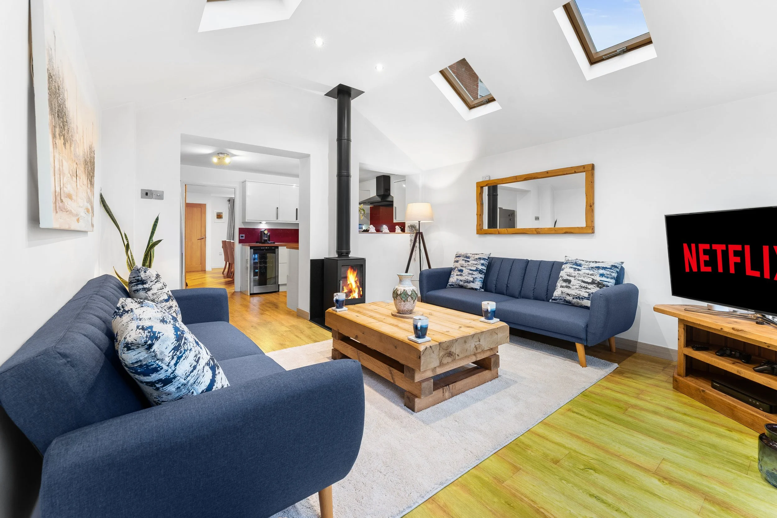Living room with blue sofas, a wooden coffee table, a fireplace with a chimney, a television displaying Netflix, and skylights in a white ceiling.