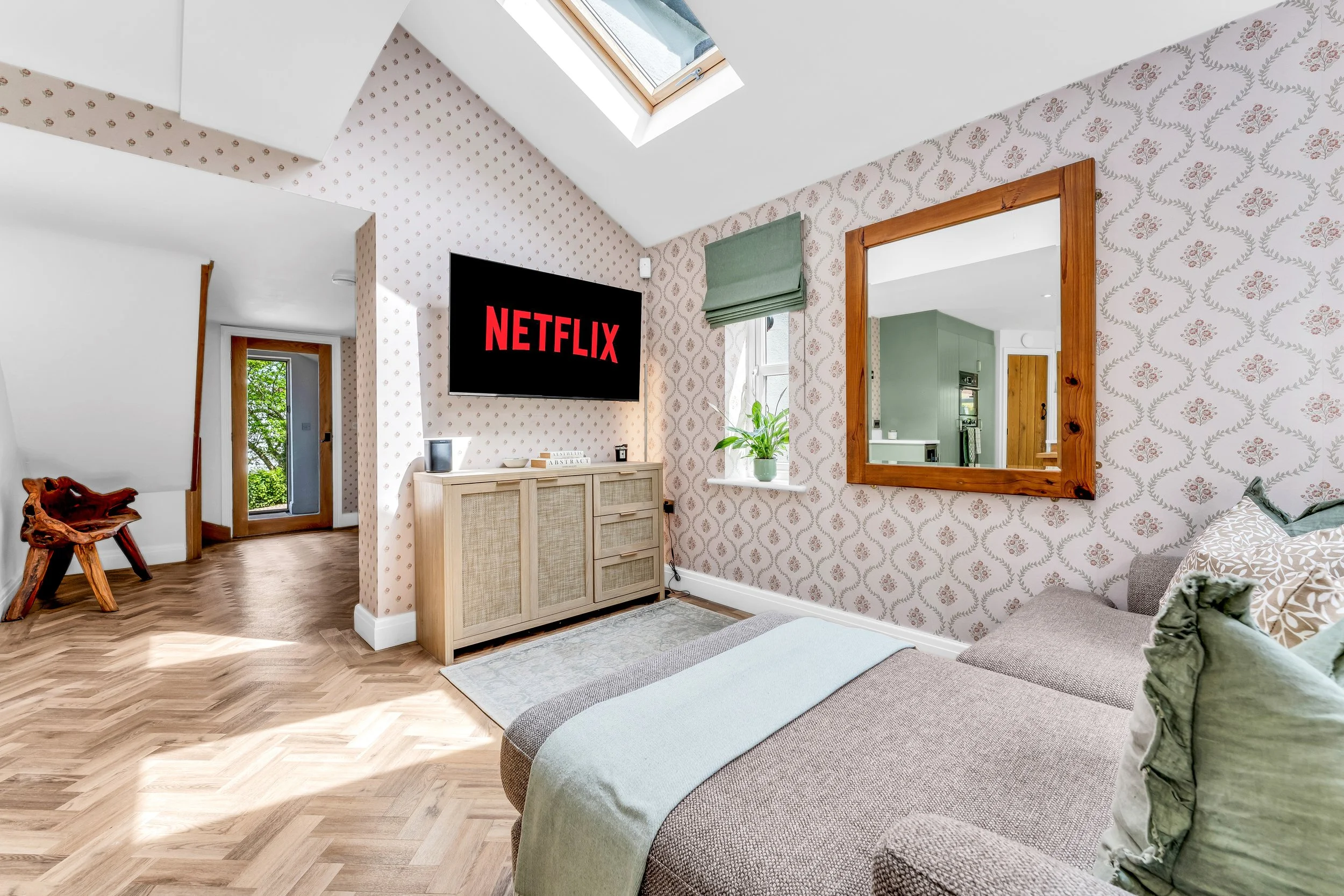 A cozy living room with patterned wallpaper, a wall-mounted Netflix TV, a wooden mirror, a bed with pillows, and a window with a green Roman shade. There is a skylight in the ceiling and a door leading outside, with natural light illuminating the spa