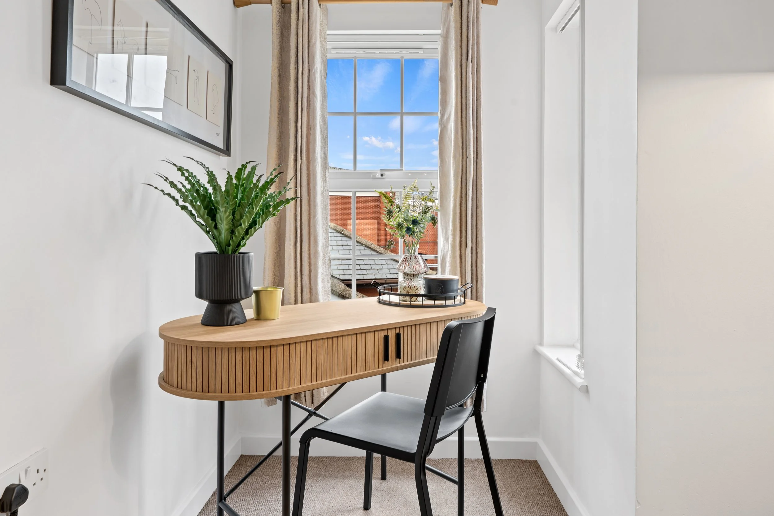 A small workspace with a wooden desk, black chair, potted plant, and a window showing a blue sky with clouds.