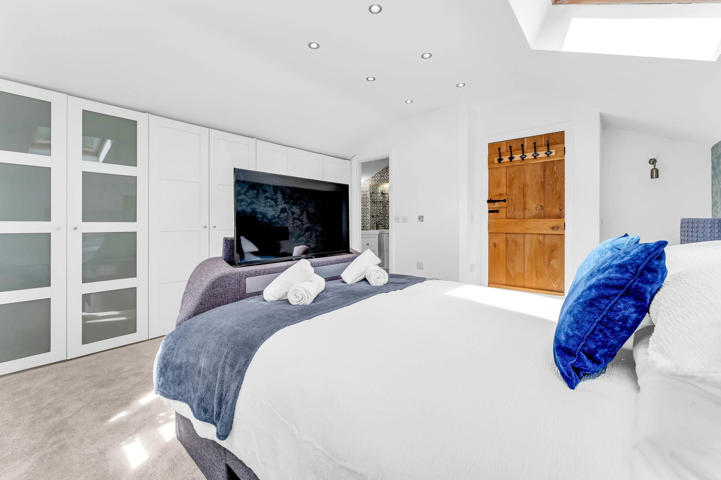 A bedroom with a bed covered in white bedding and a gray blanket, decorated with blue pillows. There are three rolled towels on the bed, a flat-screen TV on a gray upholstered stand, built-in white cabinets on one side, and a wooden door with black m