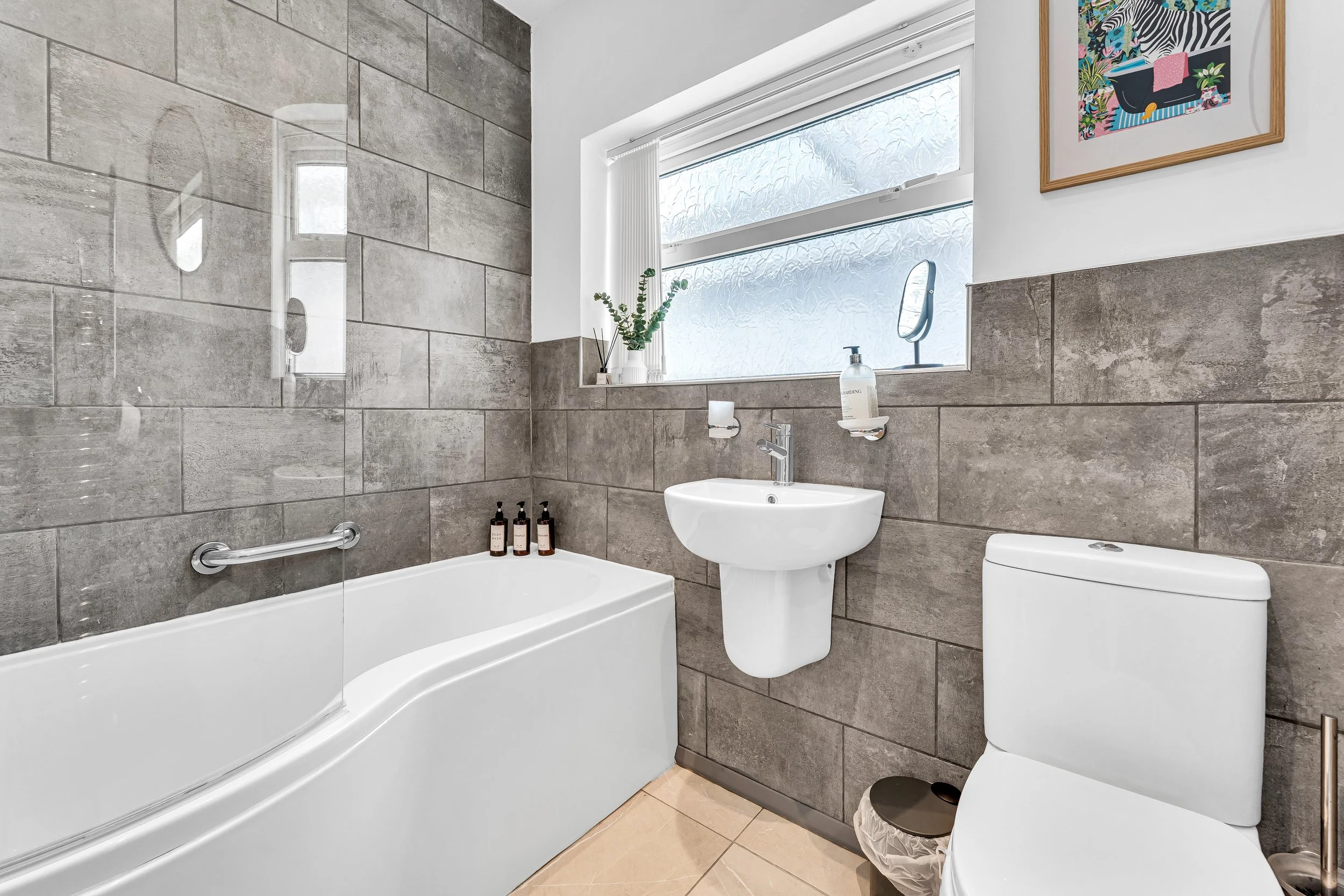 Modern bathroom with a white bathtub, small wall-mounted sink, toilet, and frosted window. Decor includes a framed colorful artwork, a small mirror, toiletries, and a potted plant.