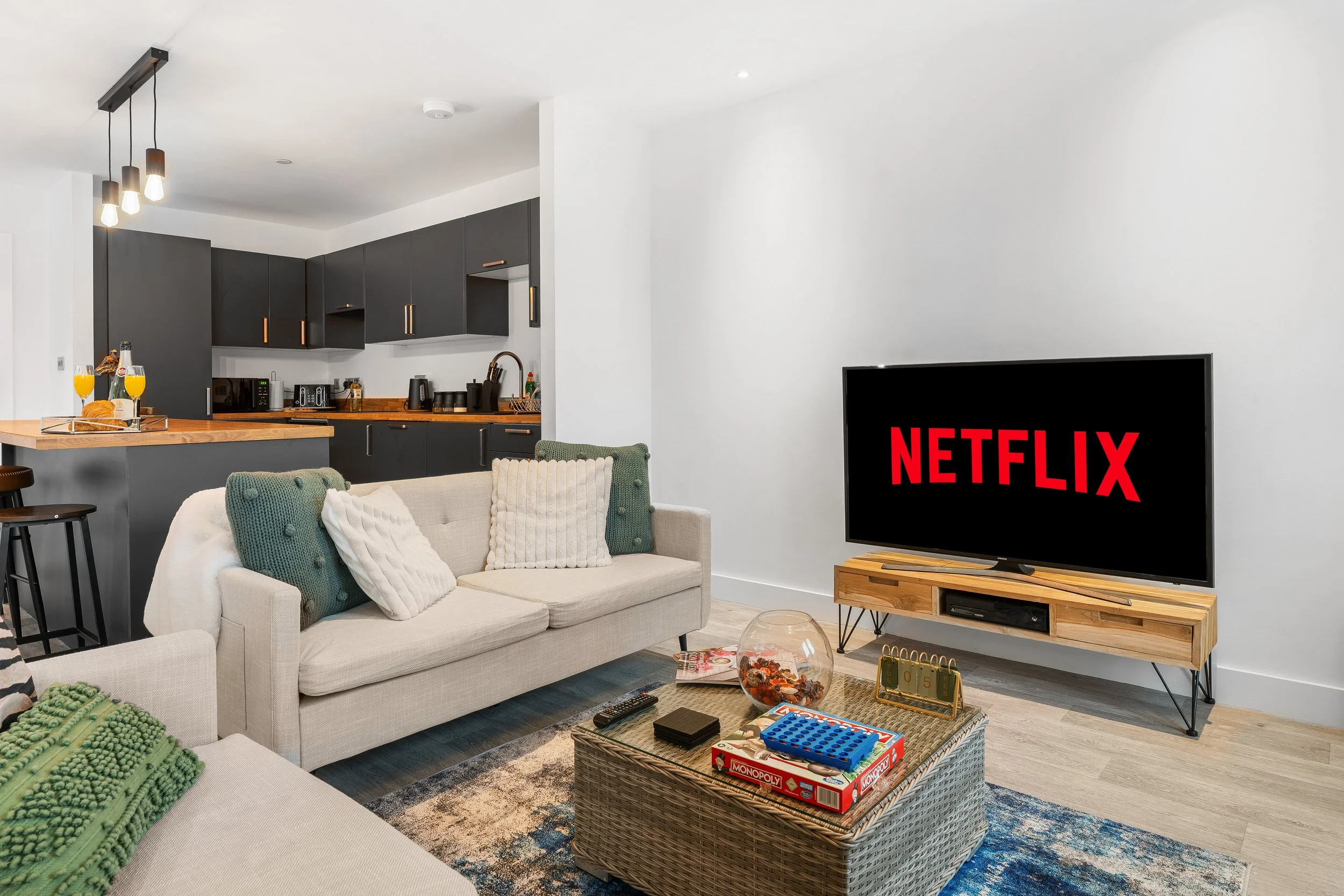 Living room with beige sofa, a wicker coffee table, and a TV displaying the Netflix logo. The background shows a kitchen with dark cabinets, an island with snacks and drinks, and some game pieces on the table.
