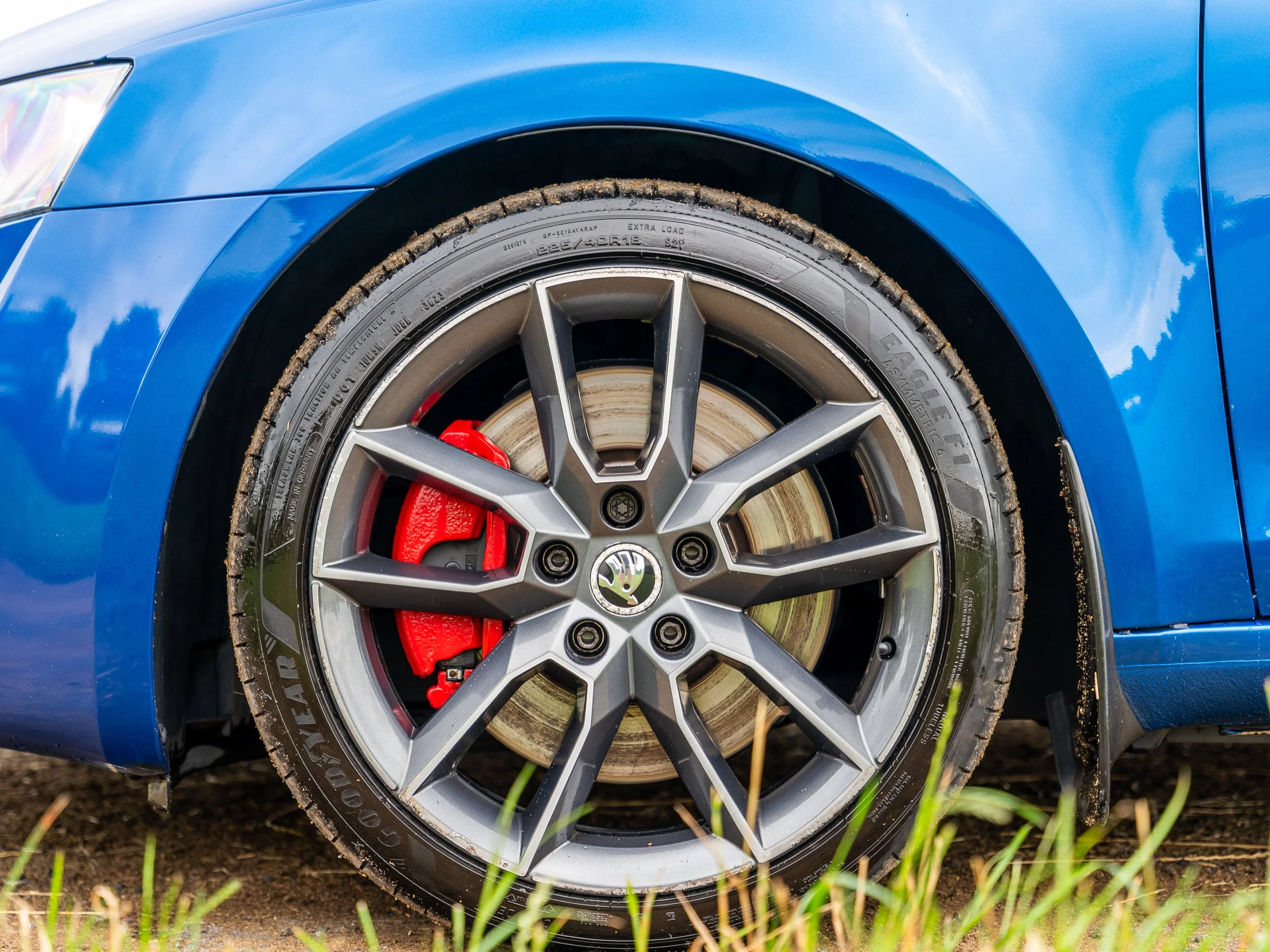Close-up of a blue car wheel with a gray alloy rim, red brake caliper, and black tire with mud around the edges, on a dirt surface with grass in the foreground.