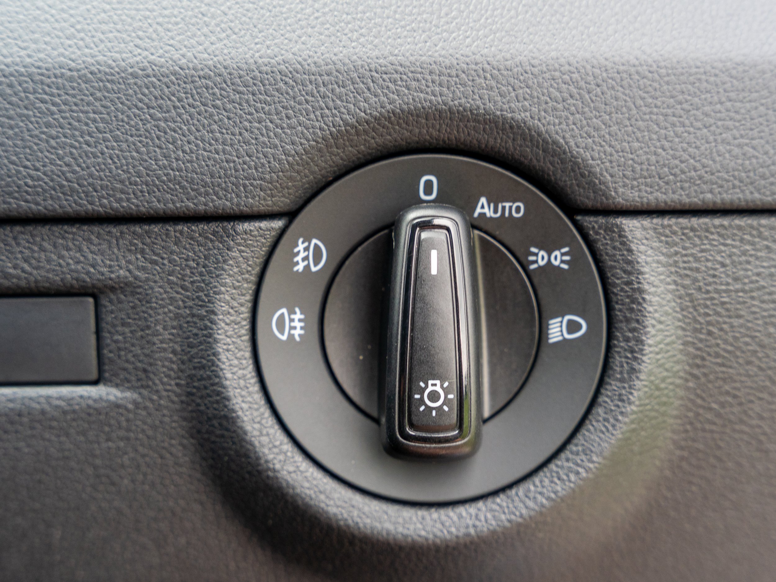 Car headlight control switch with various settings including off, auto, parking lights, headlights, and fog lights, mounted on a textured dashboard.