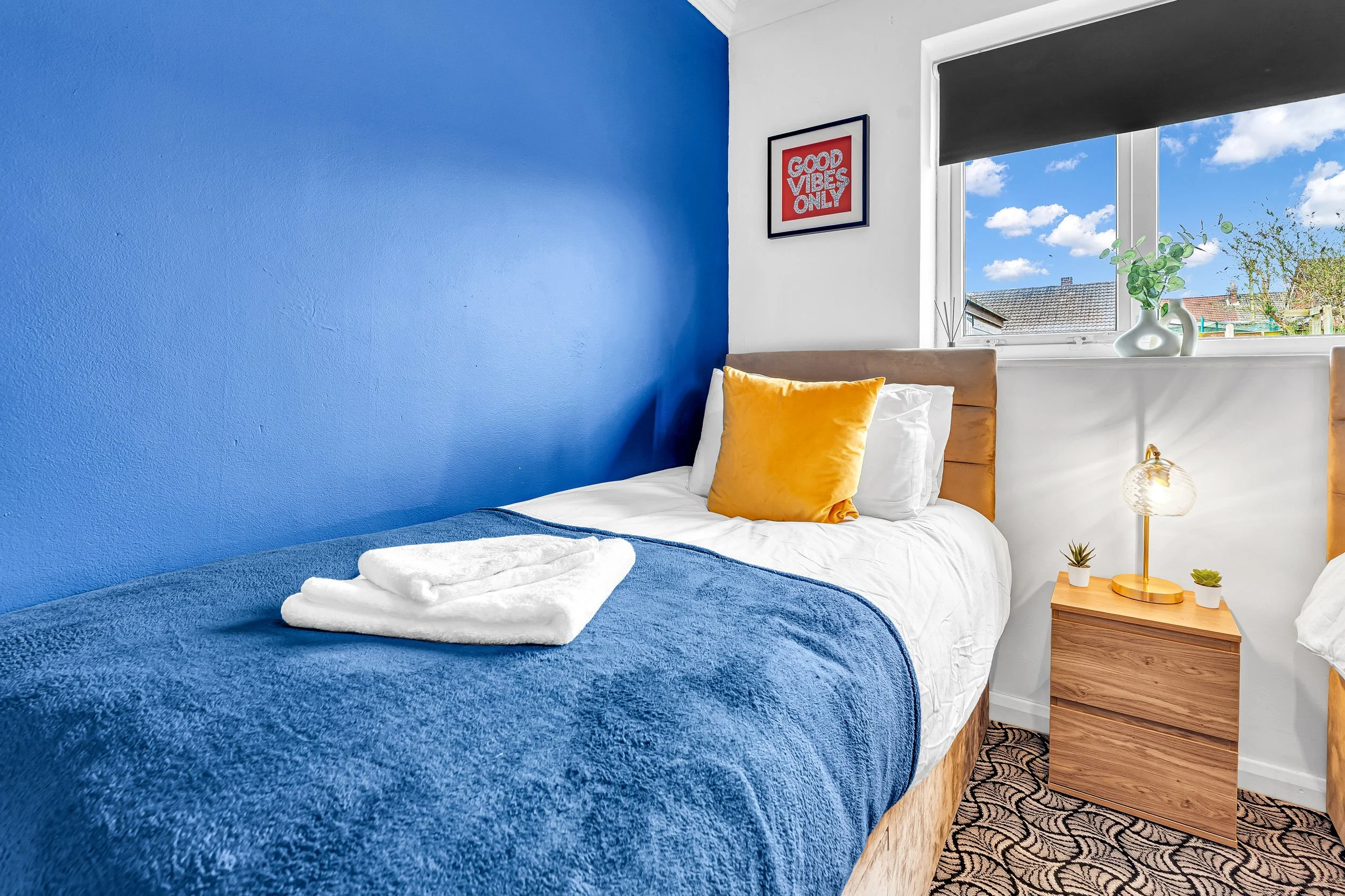 A neatly made bed with white linens, a yellow pillow, and a blue blanket, next to a nightstand with a modern lamp and small potted plants, set in a room with a blue accent wall and a window showing a view of the sky and neighboring houses.