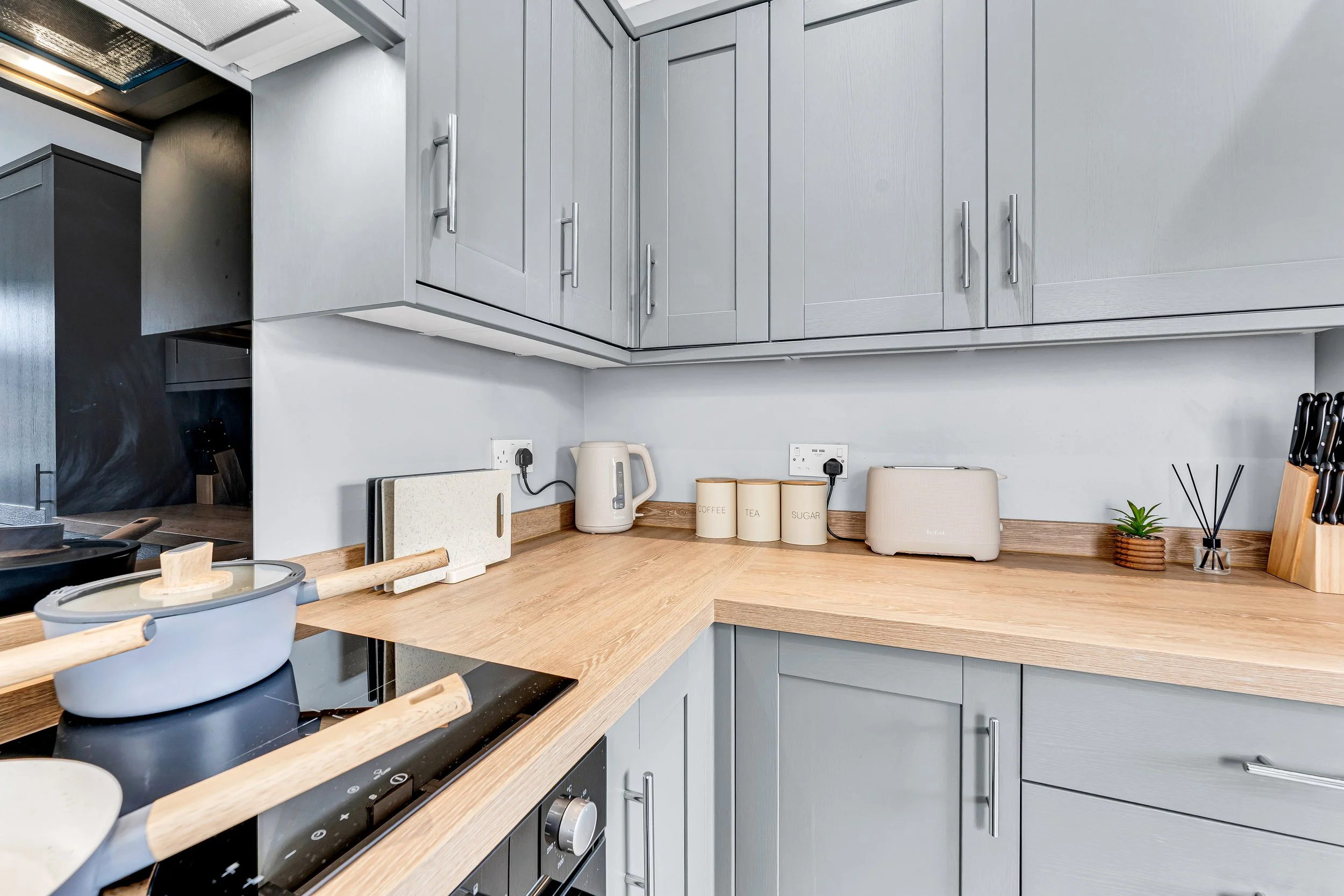 Modern kitchen with wooden countertops, gray cabinets, electric stove, white kettle, bread box, small plant, and cooking utensils on the counter.