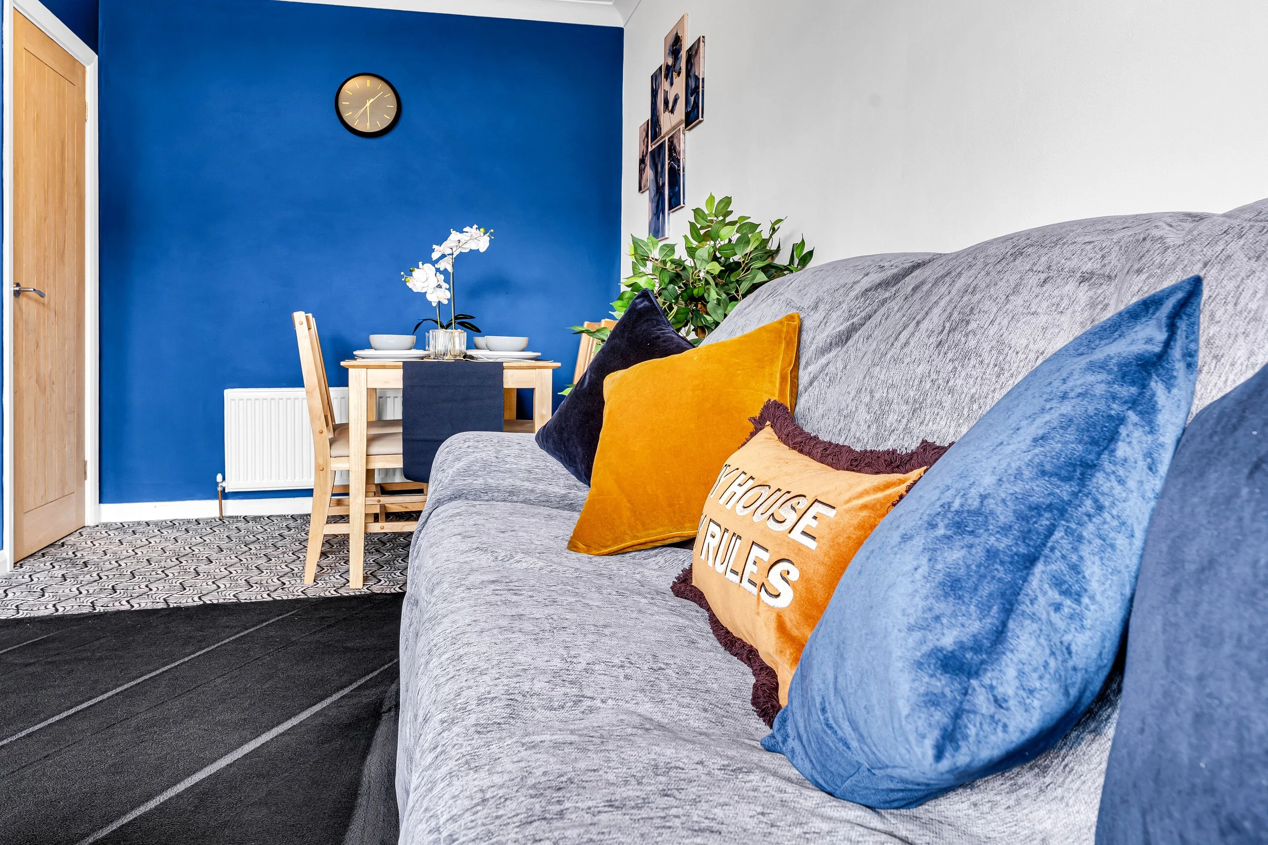 Living room with a light gray couch decorated with multiple colored pillows, including a tan pillow with text, set against a white wall. In the background, has a blue accent wall with a round clock, a small wooden dining table set with four bowls and