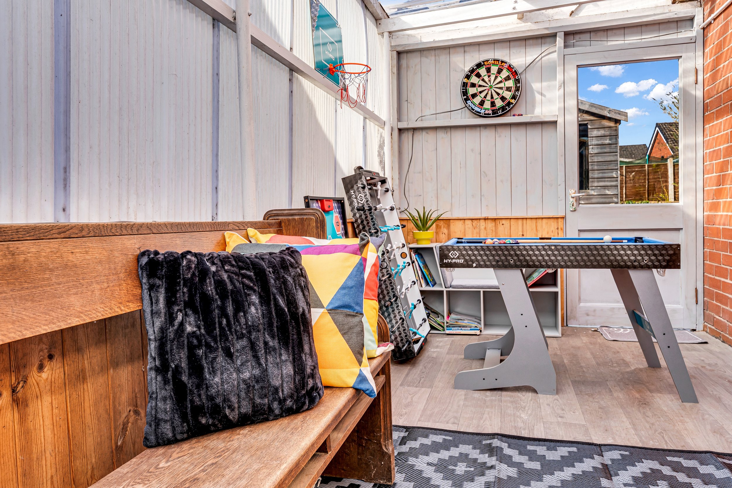 Enclosed porch with a wooden bench, black fur blanket, colorful pillow, dartboard, kid's soccer goal, bookshelf, foosball table, plant, and a glass door revealing a backyard with houses and trees.