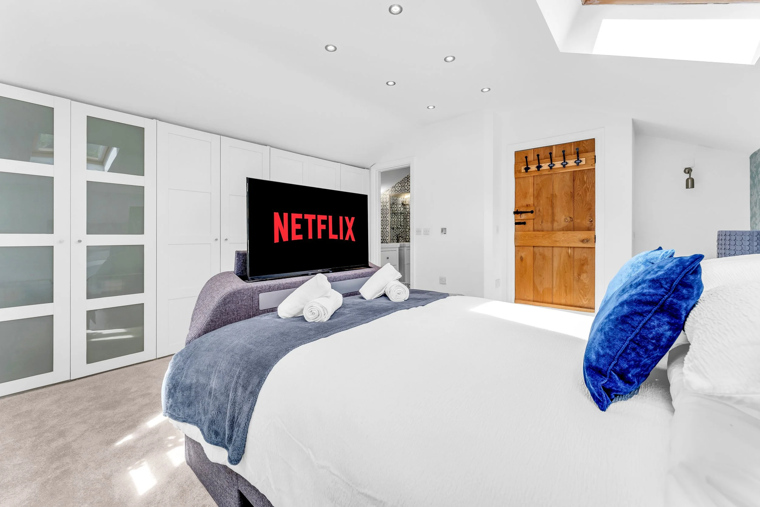 Modern bedroom with a large bed covered in white and blue bedding, two rolled white towels on the bed, a blue velvet pillow, a flat-screen TV showing the Netflix logo, a white closet with frosted glass doors, a wooden door with black hooks, a small w