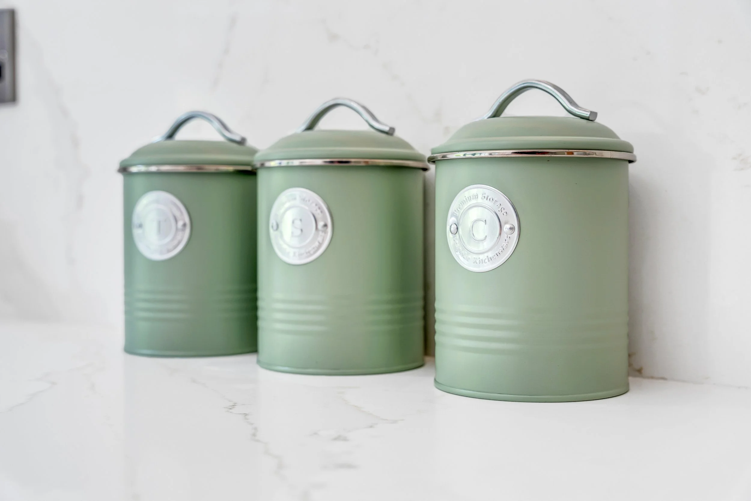 Three green storage containers with silver handles on a white marble surface against a plain white wall.