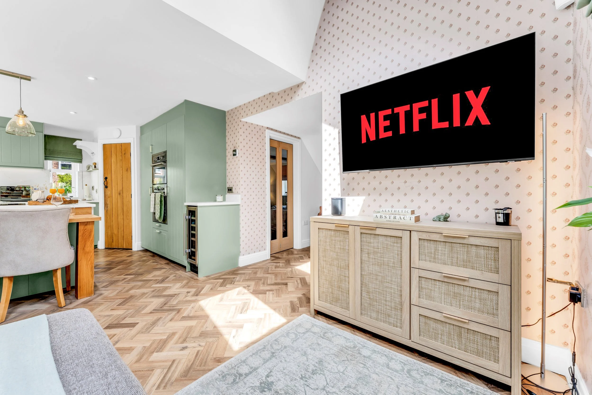 Living room with a large flat-screen TV displaying the Netflix logo on a patterned wall, a beige wooden cabinet, and light-colored wallpaper.