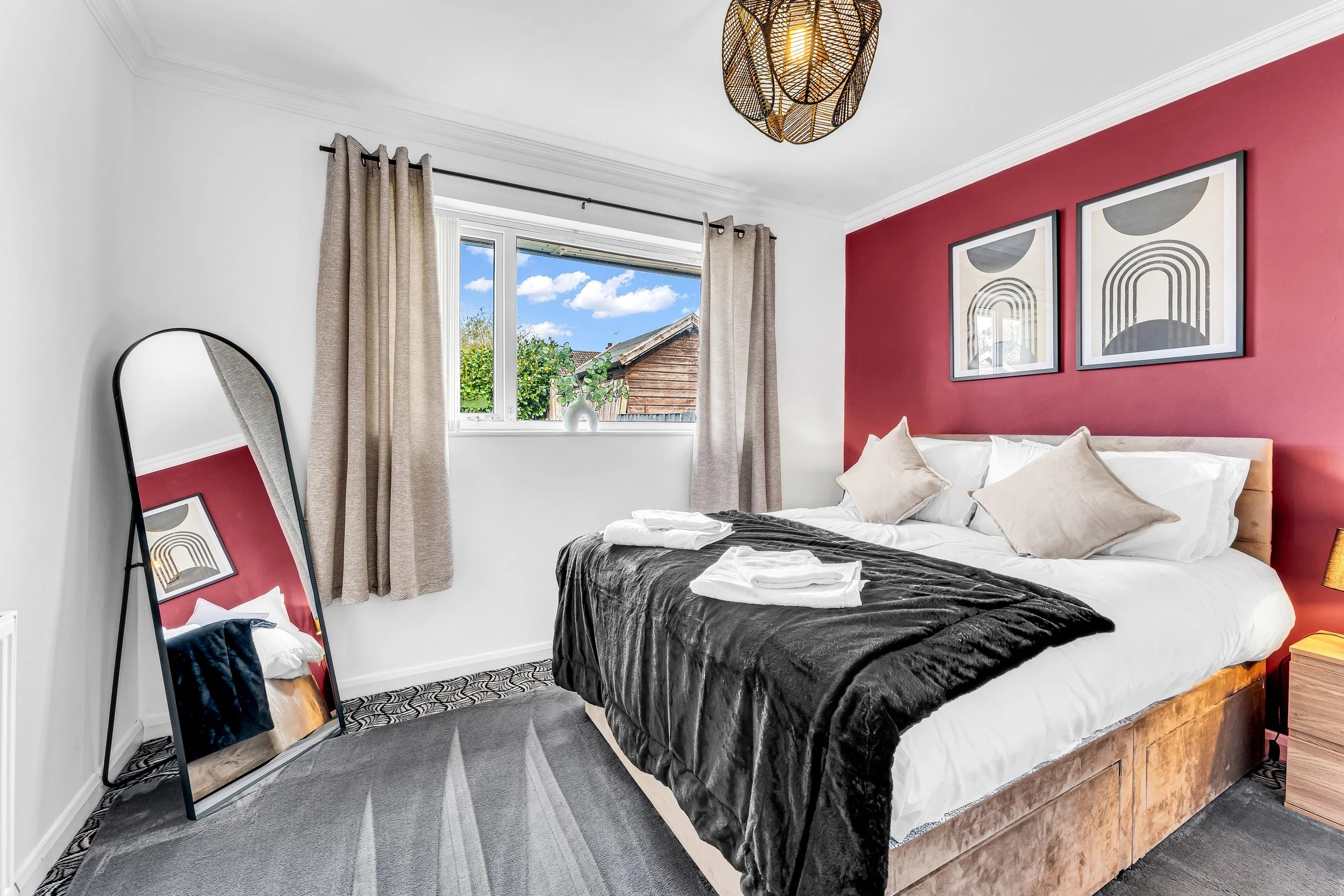 A neatly made bed with white sheets, beige pillows, and a black throw blanket in a modern bedroom with a maroon accent wall, framed artwork, and a window showing a blue sky and greenery.