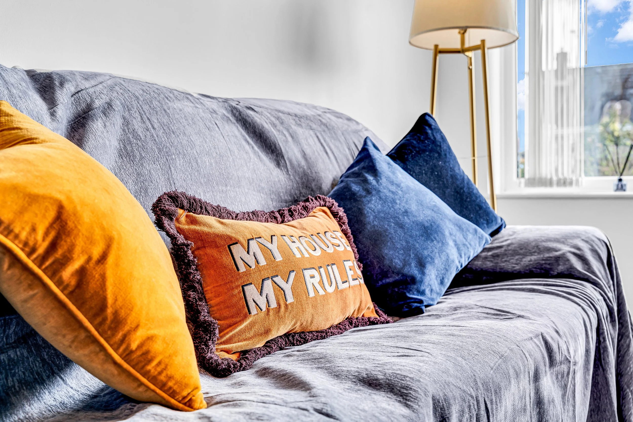 A gray sofa with orange, yellow, and blue pillows, including one with "MY HOUSE MY RULES" embroidered on it, in front of a window with white blinds and a standing lamp.