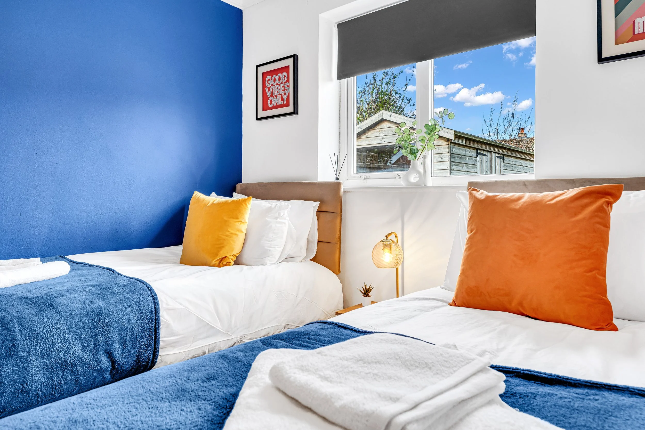A brightly lit bedroom with two beds, each with white linens, orange pillows, and blue throws. There is a window showing a backyard with a wooden shed and trees, a small lamp on the nightstand, and framed artwork on the walls, including one with the 