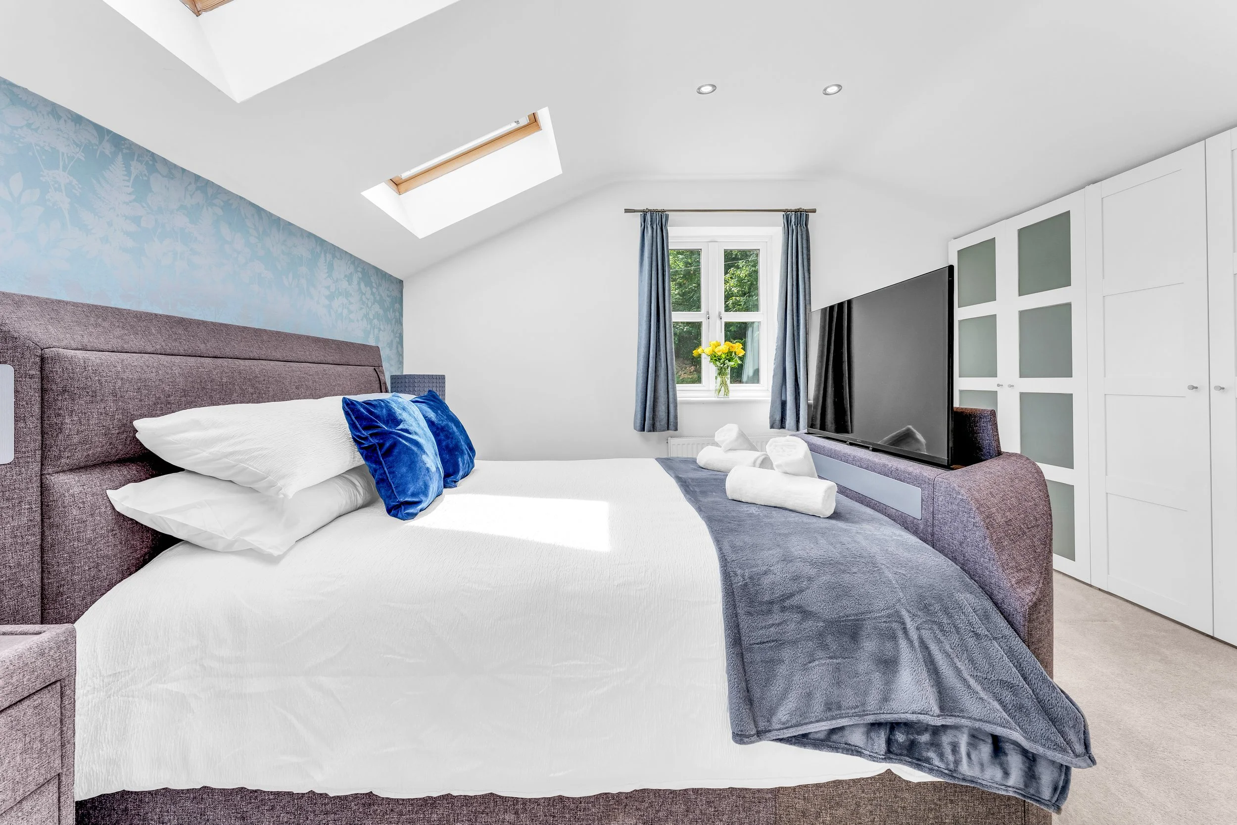 A bedroom with a sloped ceiling, a large bed with white sheets and blue pillows, a window with blue curtains and a bouquet of yellow flowers, a TV, and a white wardrobe.
