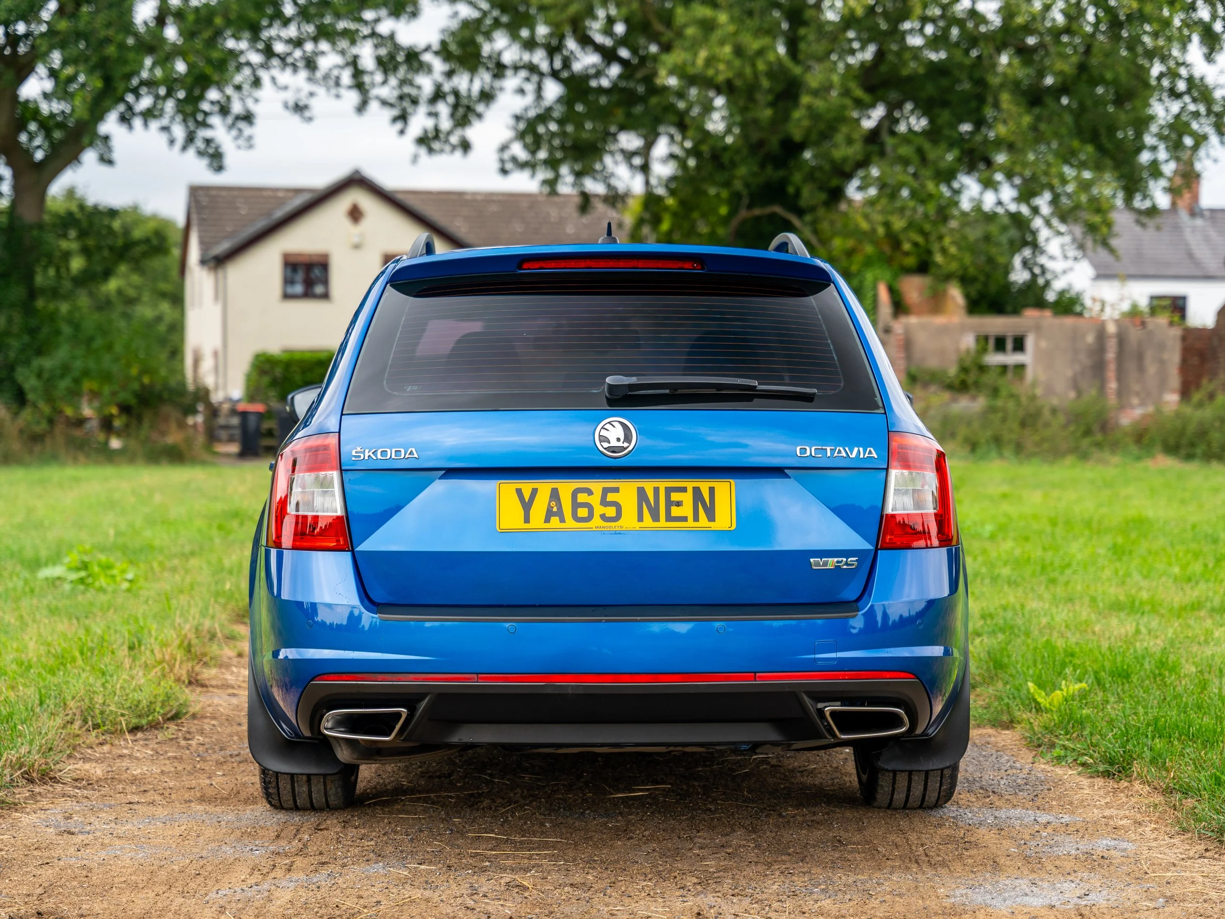 Rear view of a blue Skoda Octavia VRS parked on a dirt path with grass on either side, with houses and trees in the background.