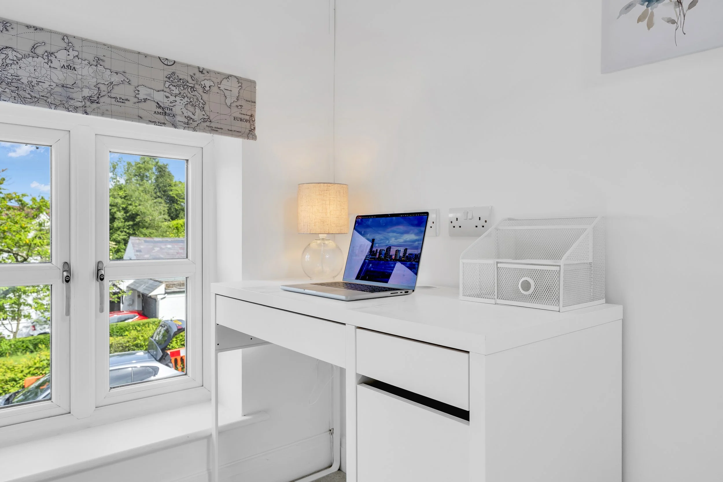 A white desk near a window with a view of trees and parked cars outside. On the desk, there is a laptop showing a city skyline, a table lamp with a fabric shade, and a white mesh pen holder. A large world map mural is mounted above the window.