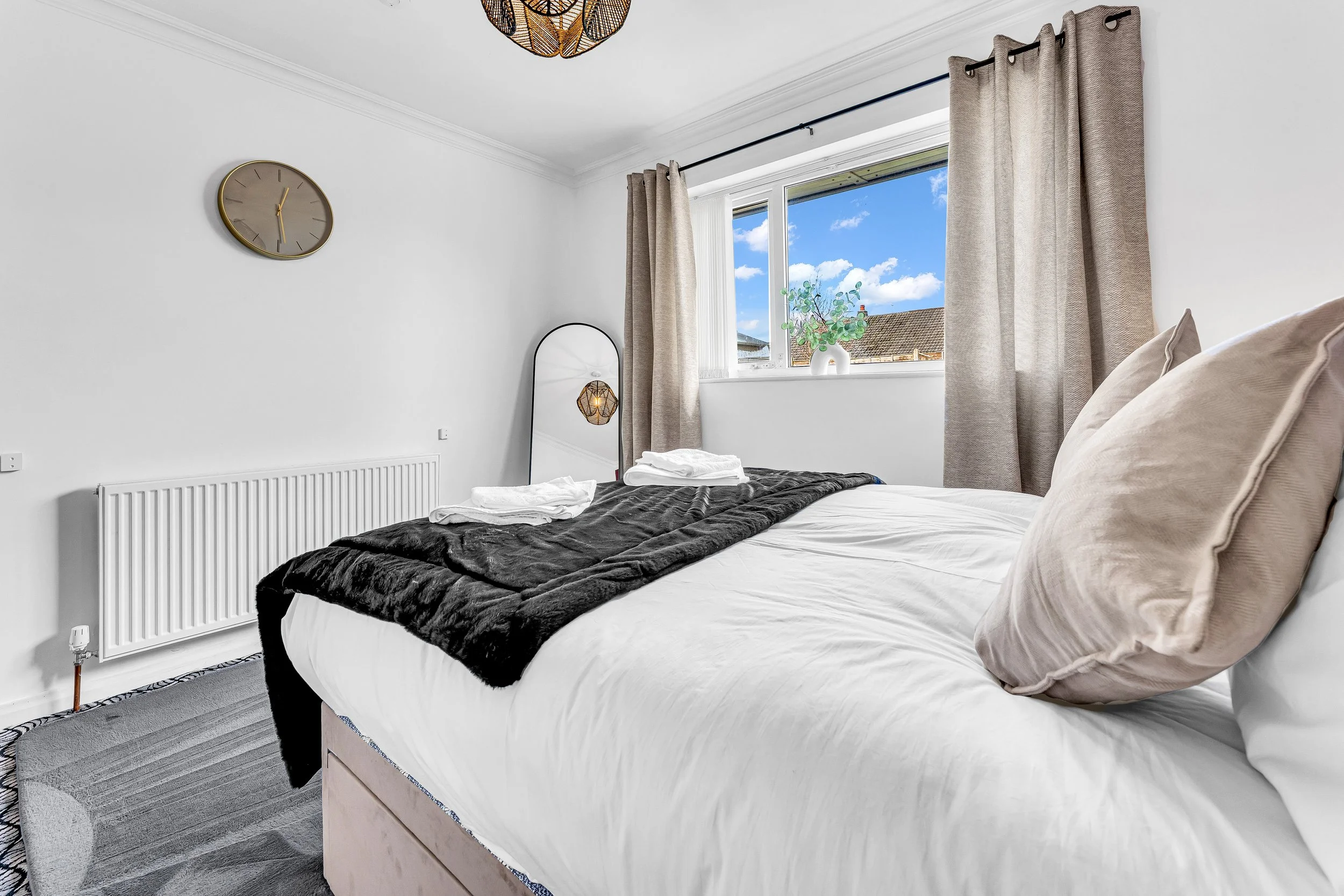 A neatly made bed with beige pillows, a black blanket, and white sheets in a bright bedroom with a window, beige curtains, a mirror, a wall clock, and a potted plant on the windowsill.