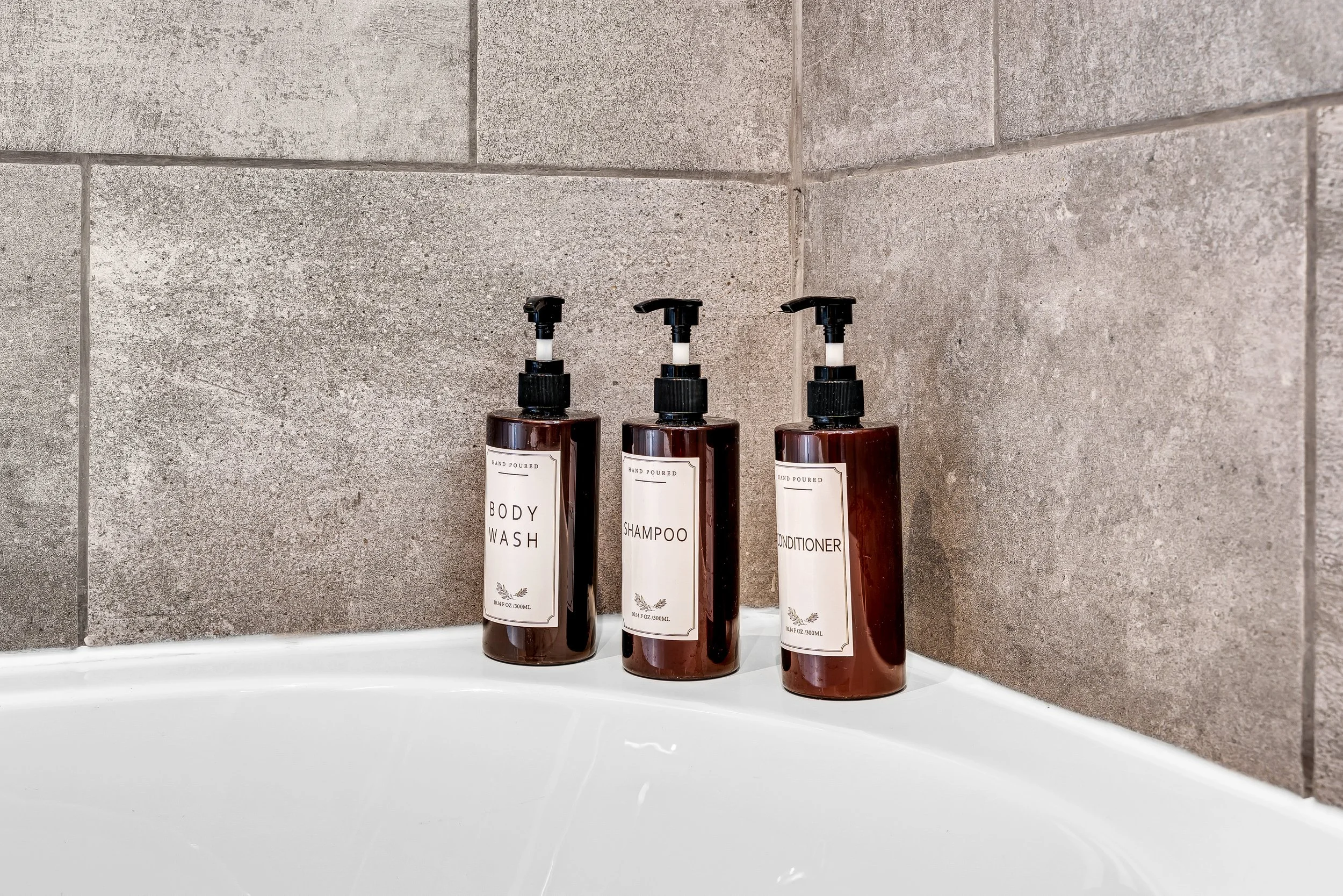 Three brown bottles with black pump dispensers labeled 'Body Wash,' 'Shampoo,' and 'Conditioner' on a white bathtub edge against a tiled wall.
