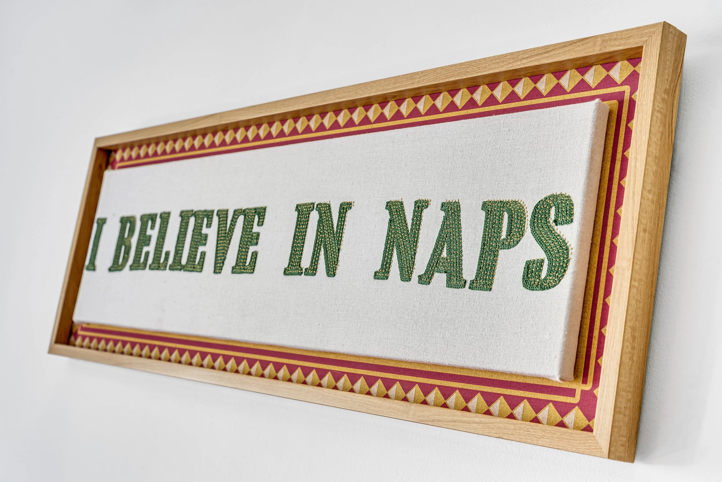 A framed fabric wall art with the text 'I BELIEVE IN NAPS' in green letters, hung on a white wall.