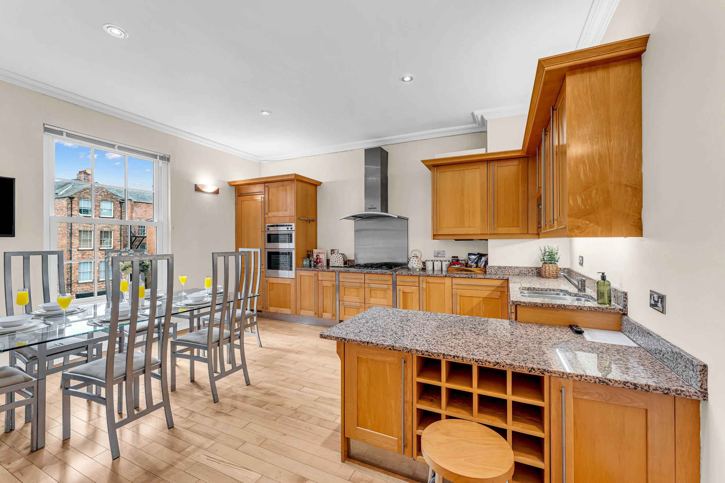 Kitchen with wooden cabinets, granite countertops, a dining table set with white plates and yellow drinks, large window showing neighboring brick buildings, and stainless steel appliances.
