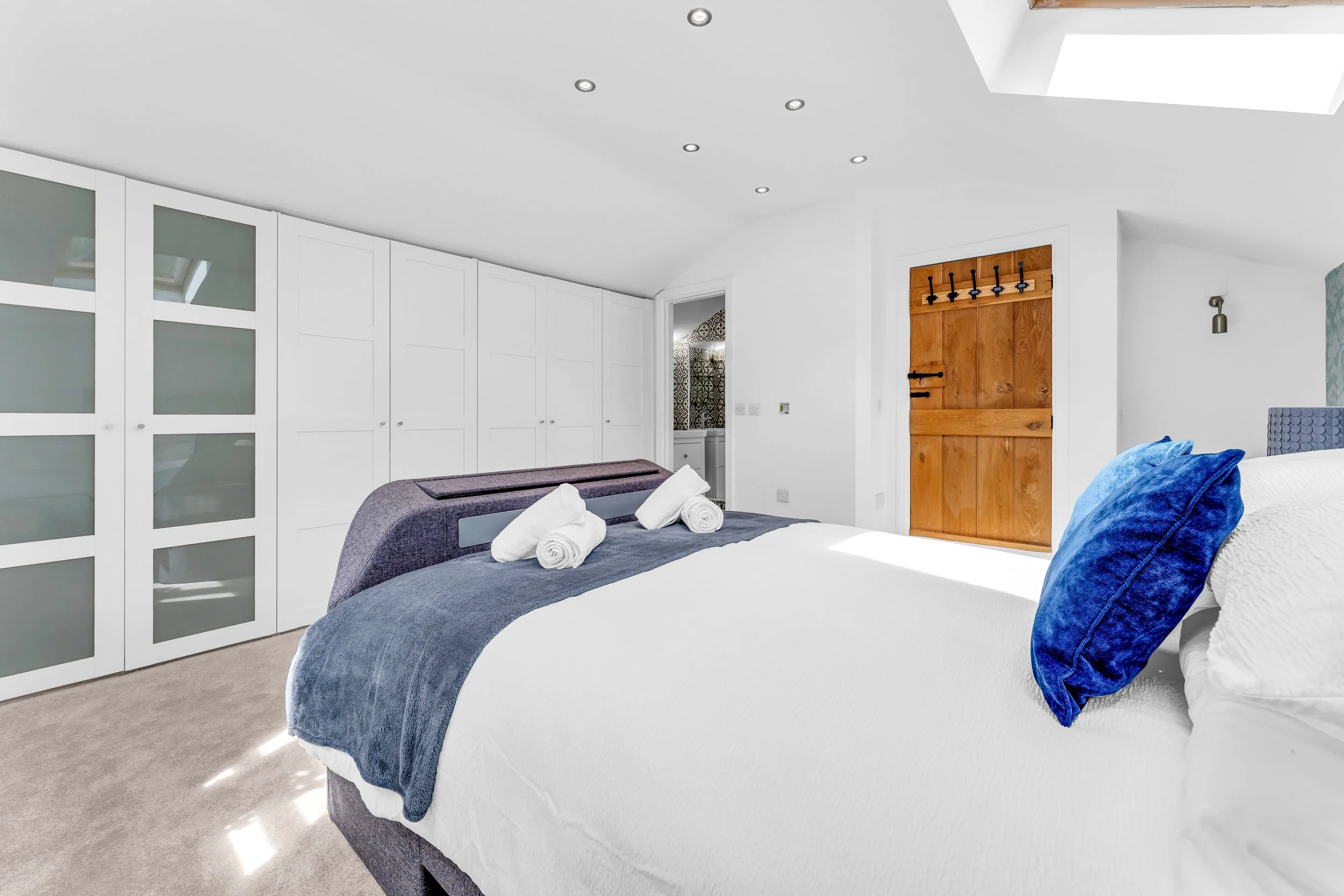 A bedroom with a bed covered in white bedding and a blue throw pillow, fresh towels on the bed, white built-in closets, a wooden door, and a small vanity room visible through an open door. The room has ceiling lights and natural light on the carpeted