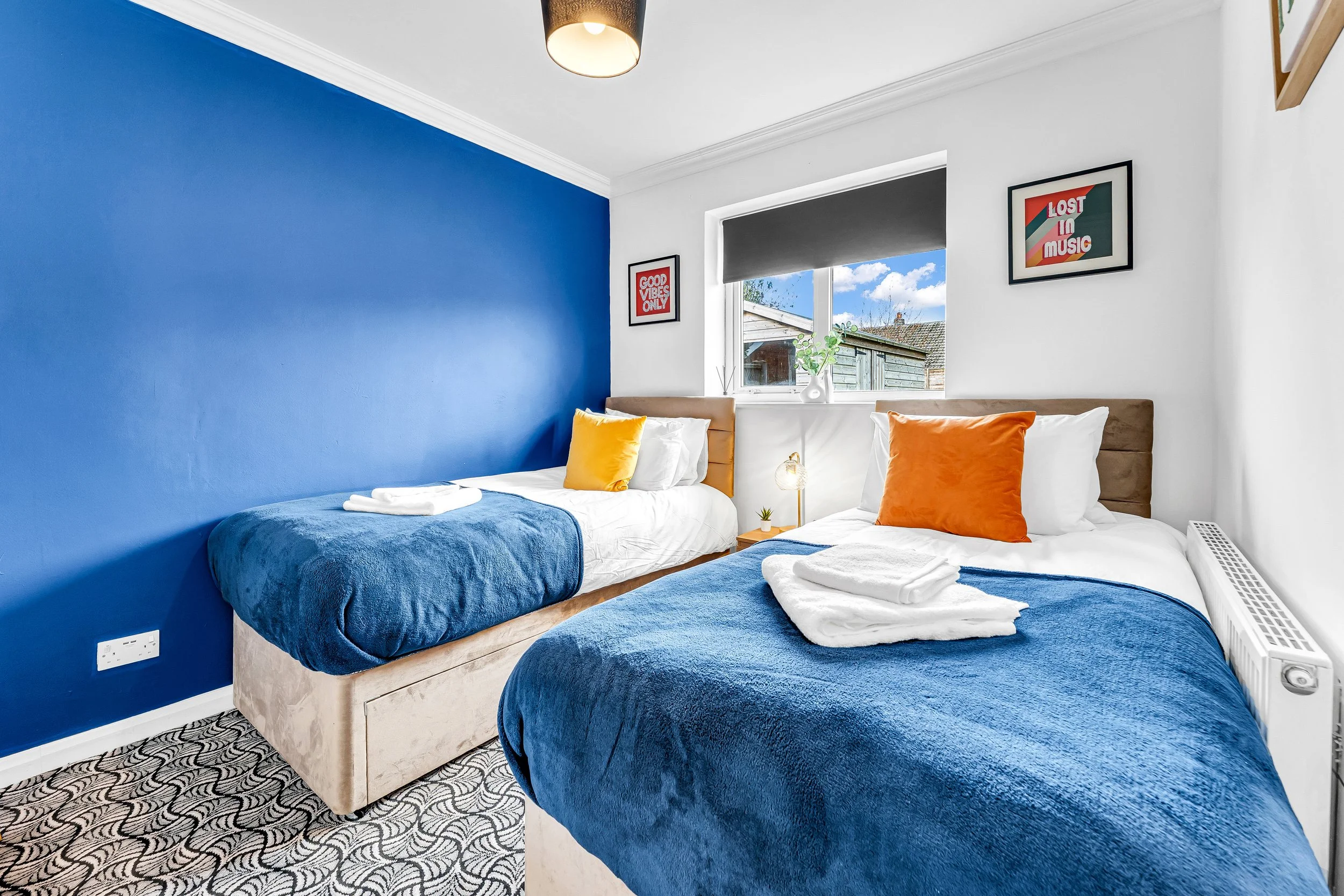 Bedroom with two single beds with blue bedspreads, one with yellow pillow and the other with orange pillow, white walls with framed artwork, window with black blind showing blue sky and clouds, patterned carpet, small bedside table with lamp and plan