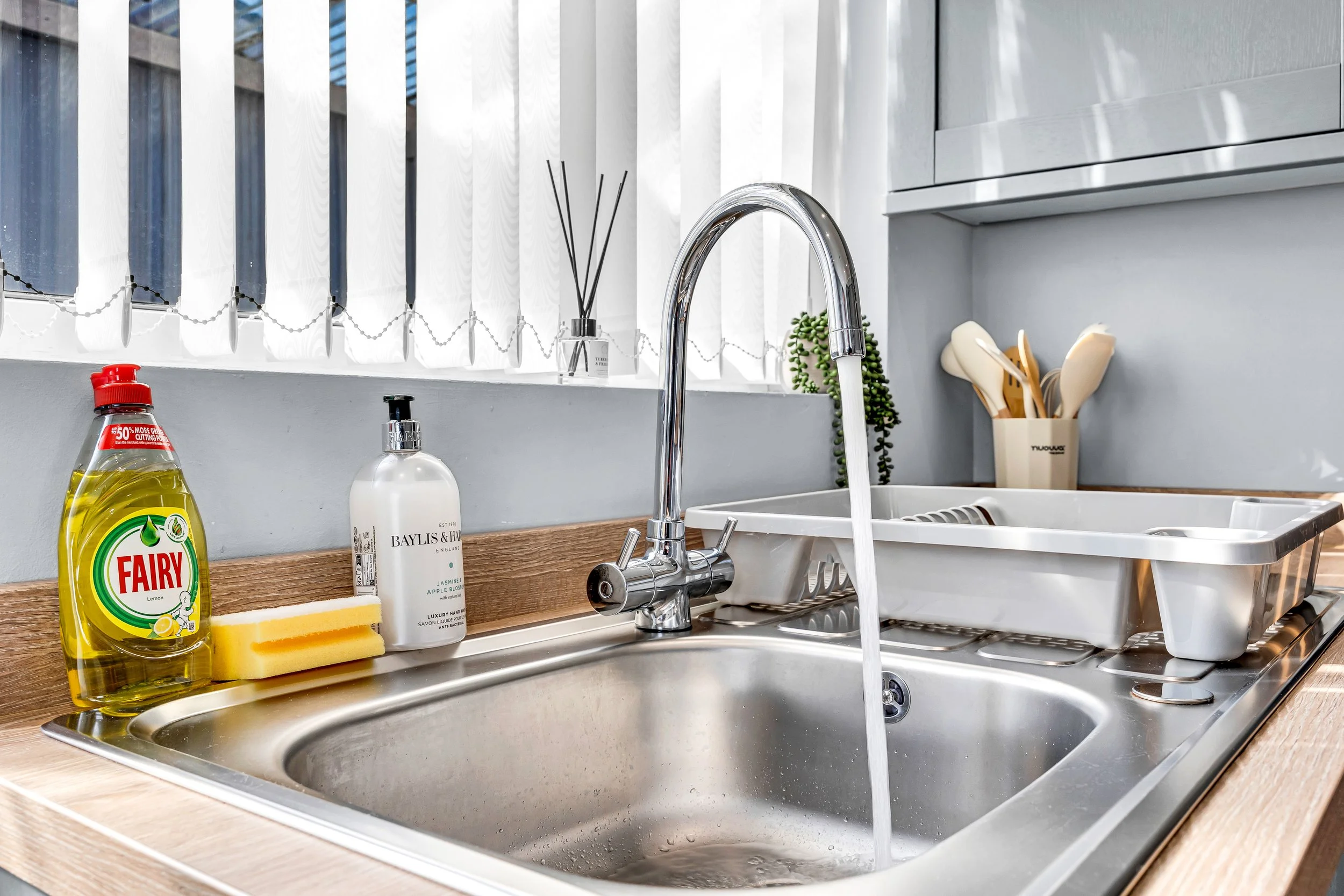Kitchen sink with running water, dish soap, sponge, bottle of hand soap, and utensils in a cup, with window blinds in the background.