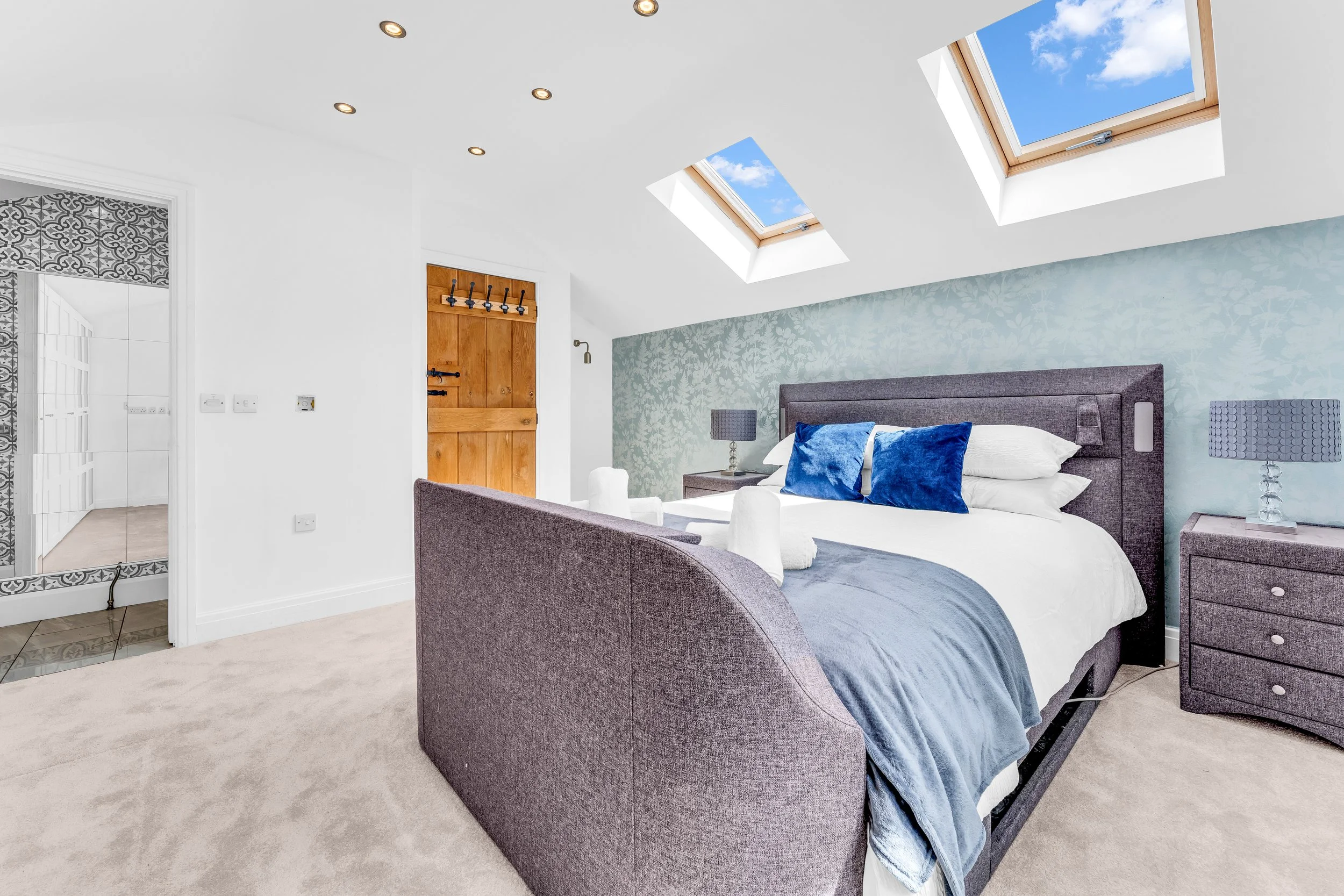 A bedroom with a gray upholstered bed, blue pillows, and a light gray bedspread. Two nightstands with matching lamps flank the bed. Two skylights illuminate the room with natural light, showing a blue sky with white clouds. There's a wooden door with