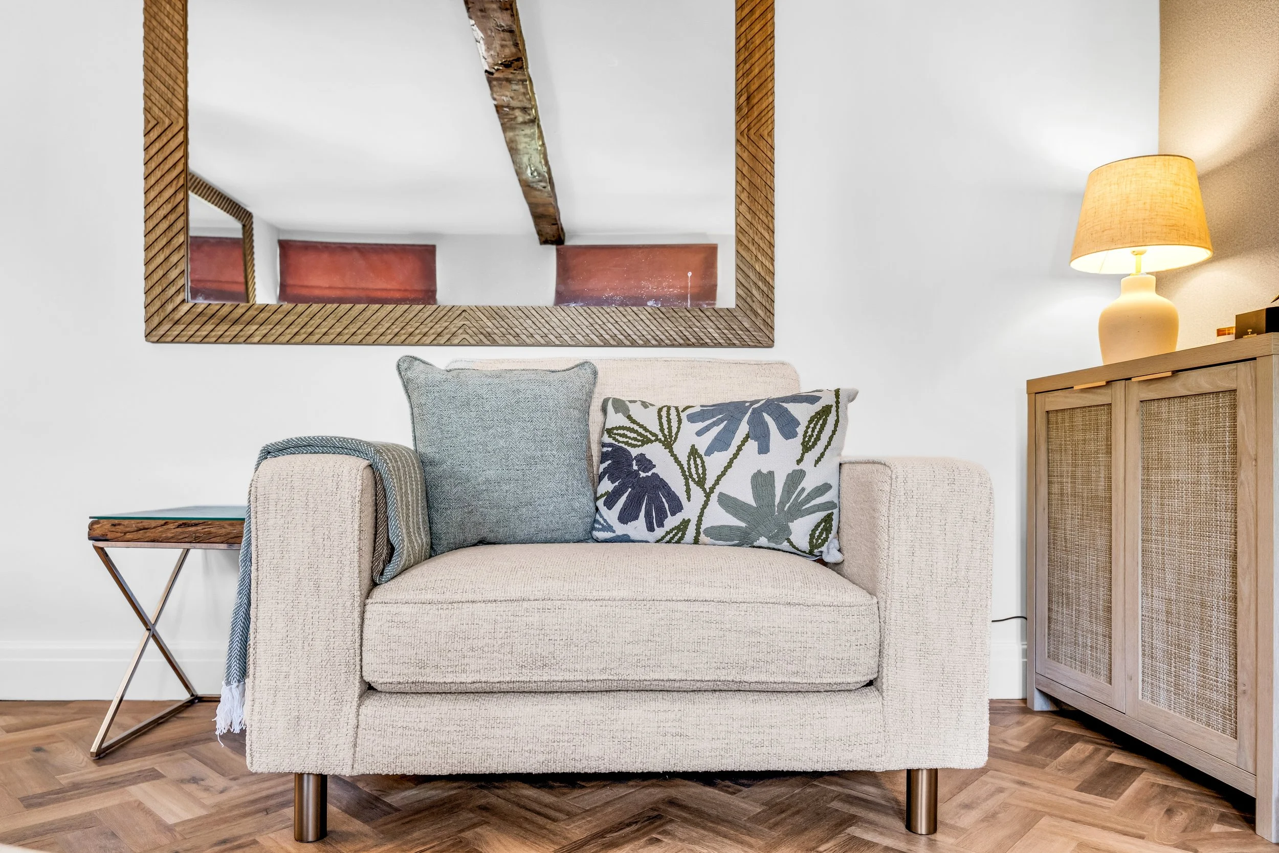 A cozy living room corner featuring a beige upholstered sofa with decorative pillows, a side table with a throw blanket, a wooden cabinet with rattan panels, a mirror with a wooden frame, a yellow table lamp, and a wooden ceiling beam, all with hardw