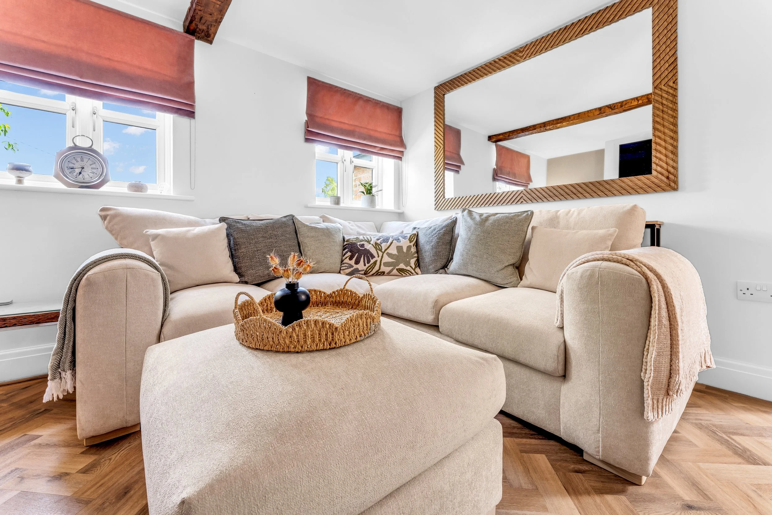 Living room with beige sectional sofa, decorative pillows, basket with black vase, wooden floor, white walls, windows with pink Roman shades, large mirror, and wooden ceiling beams.