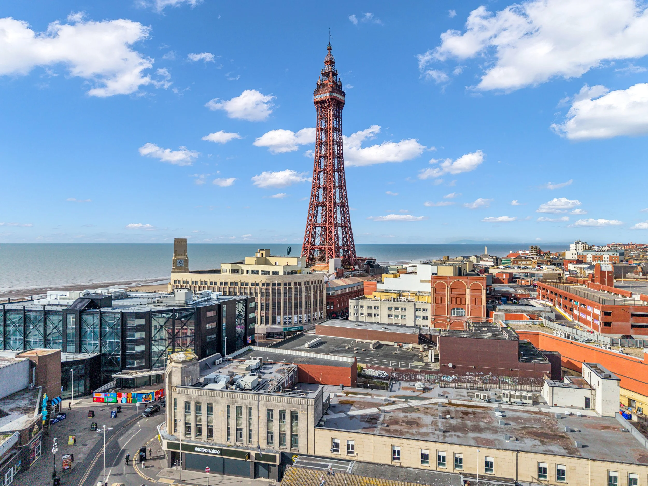 A city view with a tall iron tower near the coastline, buildings in the foreground, and a partly cloudy sky in the background.