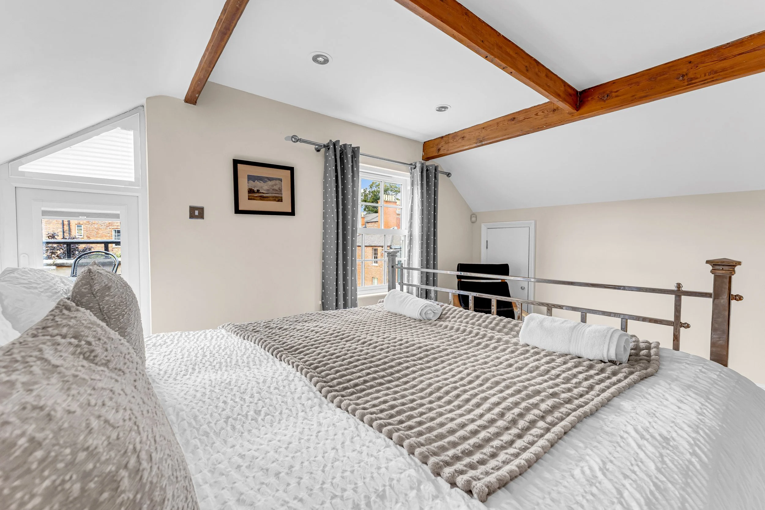 A cozy bedroom with a bed covered in white and beige textured blankets, with rolled white towels placed on top. The room has a sloped ceiling with exposed wooden beams and a large window with polka dot curtains. Outside the window, a balcony with a chair and neighboring brick buildings are visible.