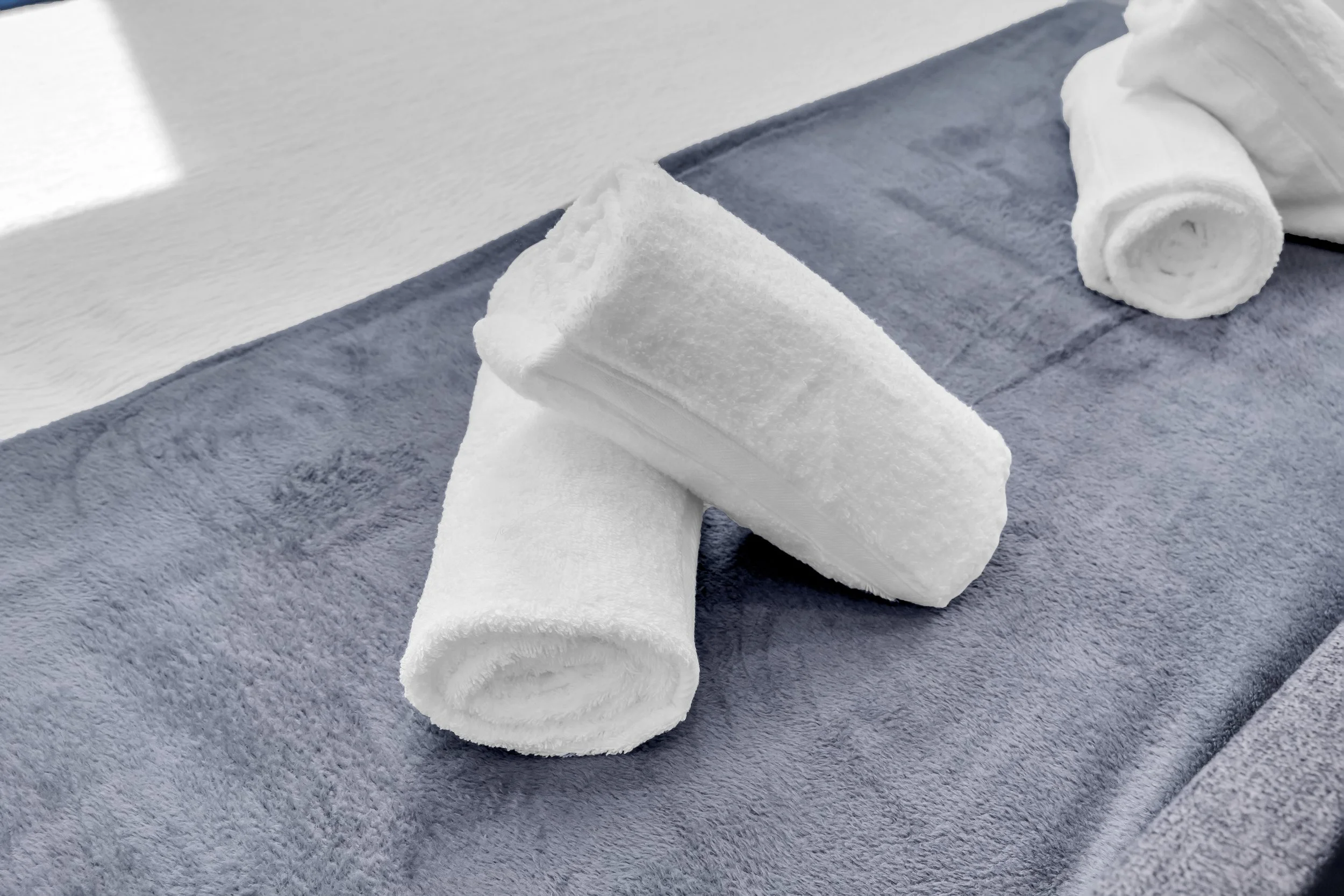 A white rolled-up towel and a folded white towel on a dark blue towel, placed on a textured gray surface next to a white wall.