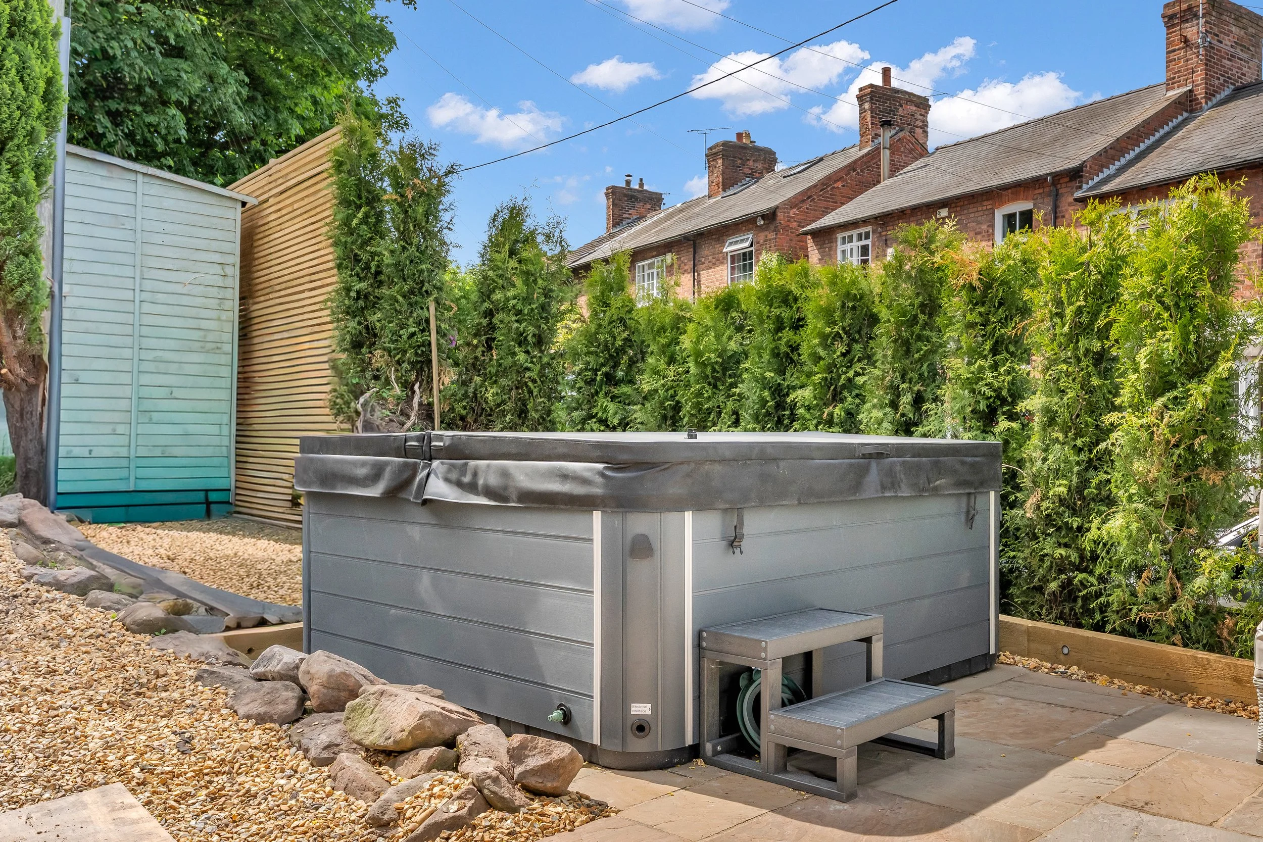 Backyard with a hot tub, gravel pathway, wooden privacy screen, and tall green bushes, with brick houses and blue sky in the background.