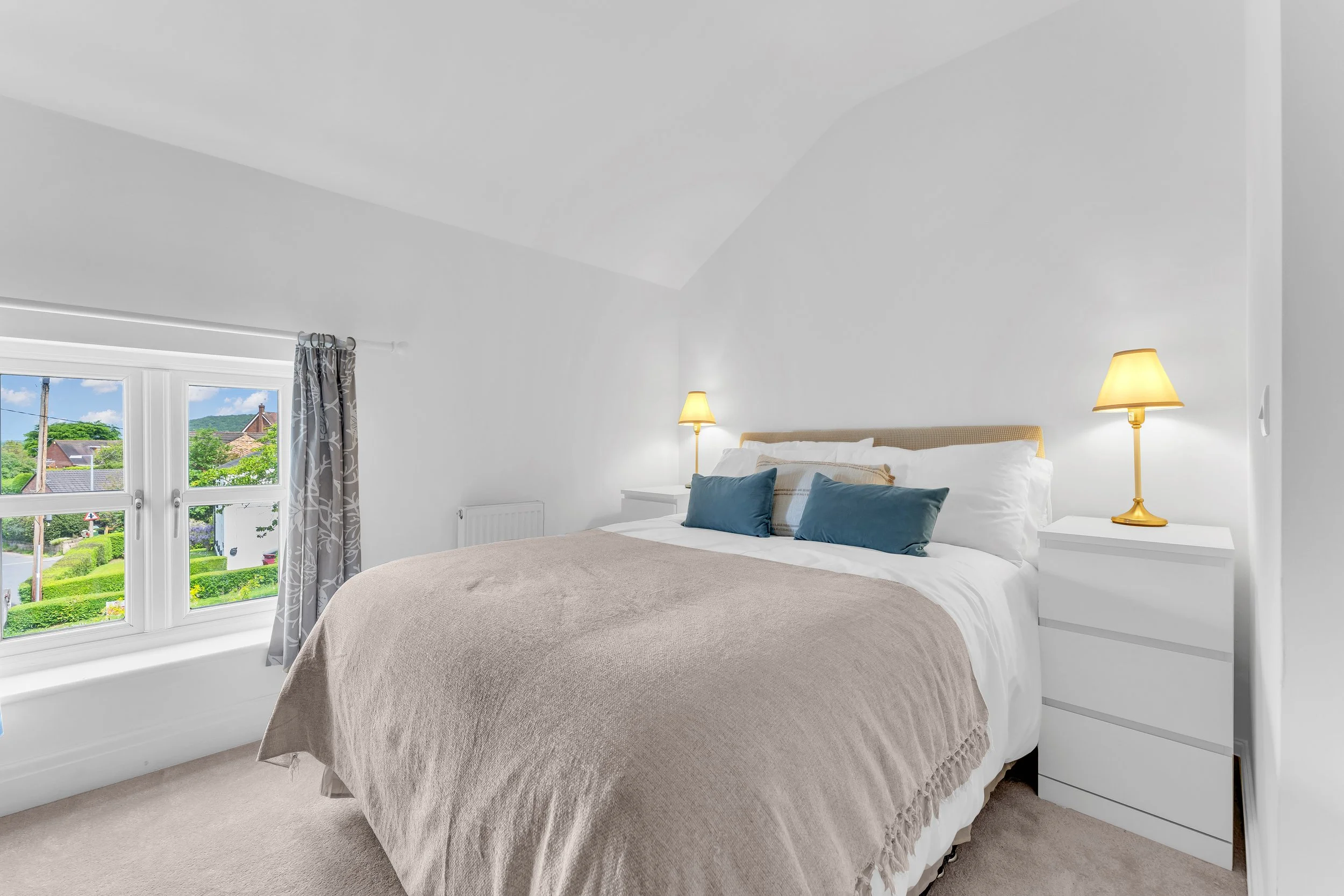 A bedroom with a large bed covered in beige bedding and multiple pillows, situated next to a white nightstand with a yellow lamp. There are two windows with views of houses, trees, and a hill outside, and curtains with a leaf pattern.