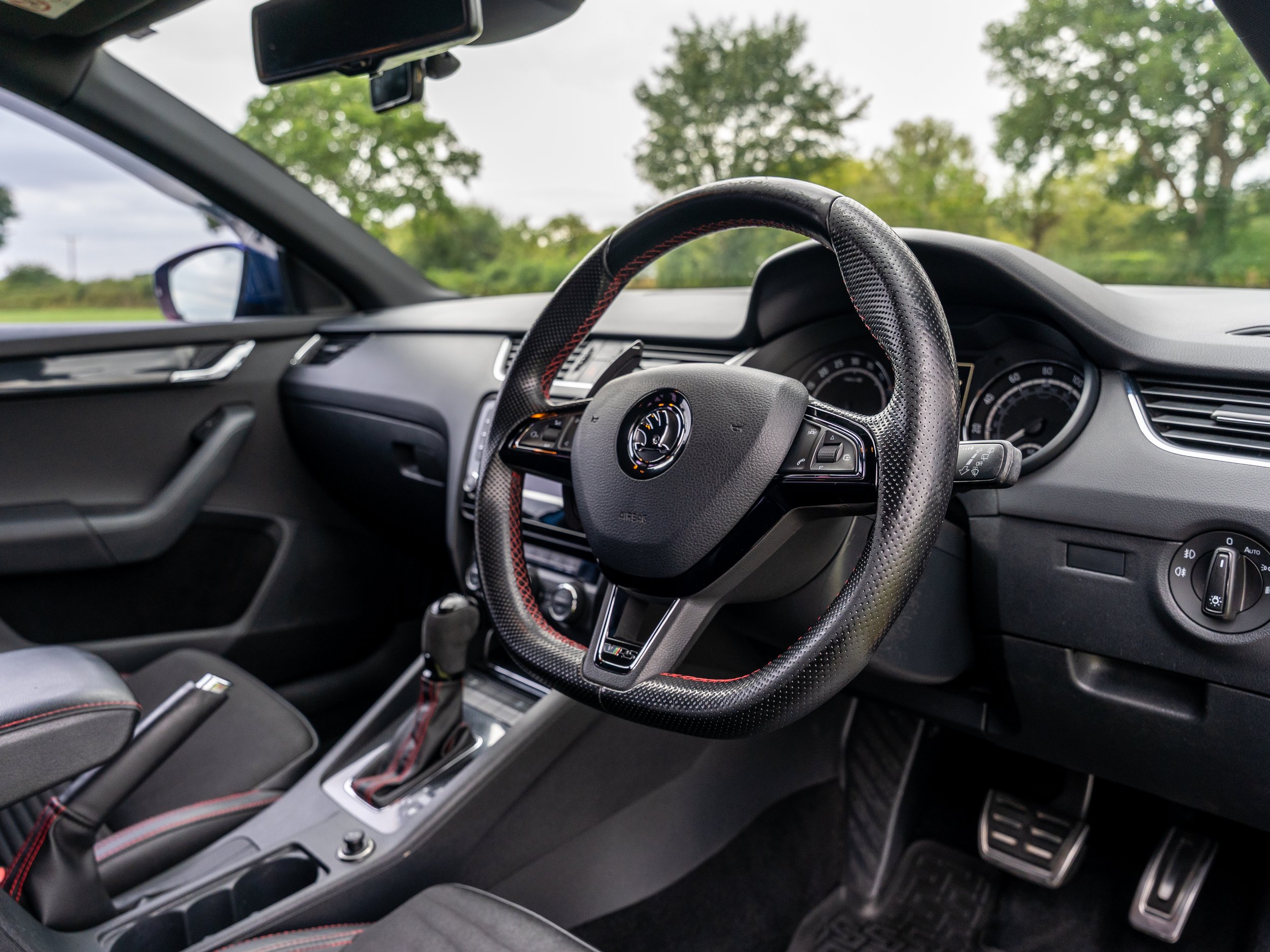 Interior of a car showing the steering wheel, dashboard, gear shift, and driver's side window with a view of trees outside.